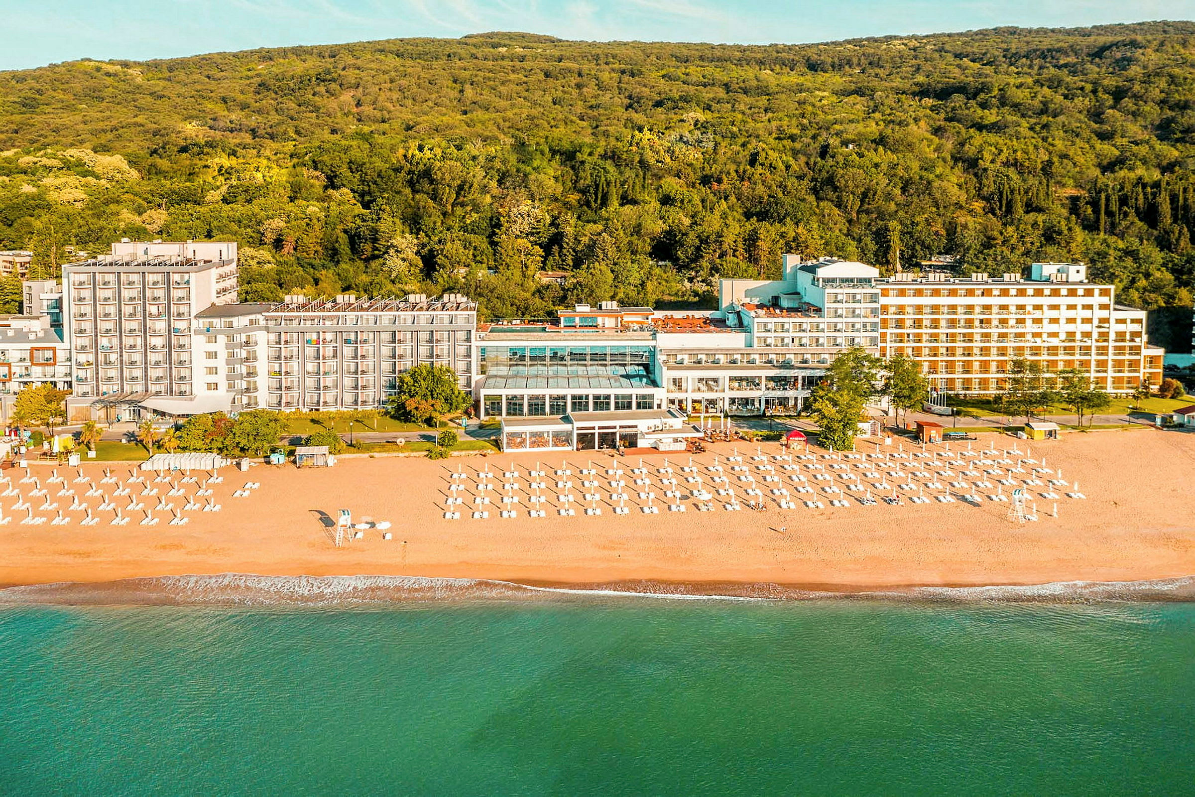 Hotell vid stranden med solstolar och parasoller framför en skog. (AI-genererad text)