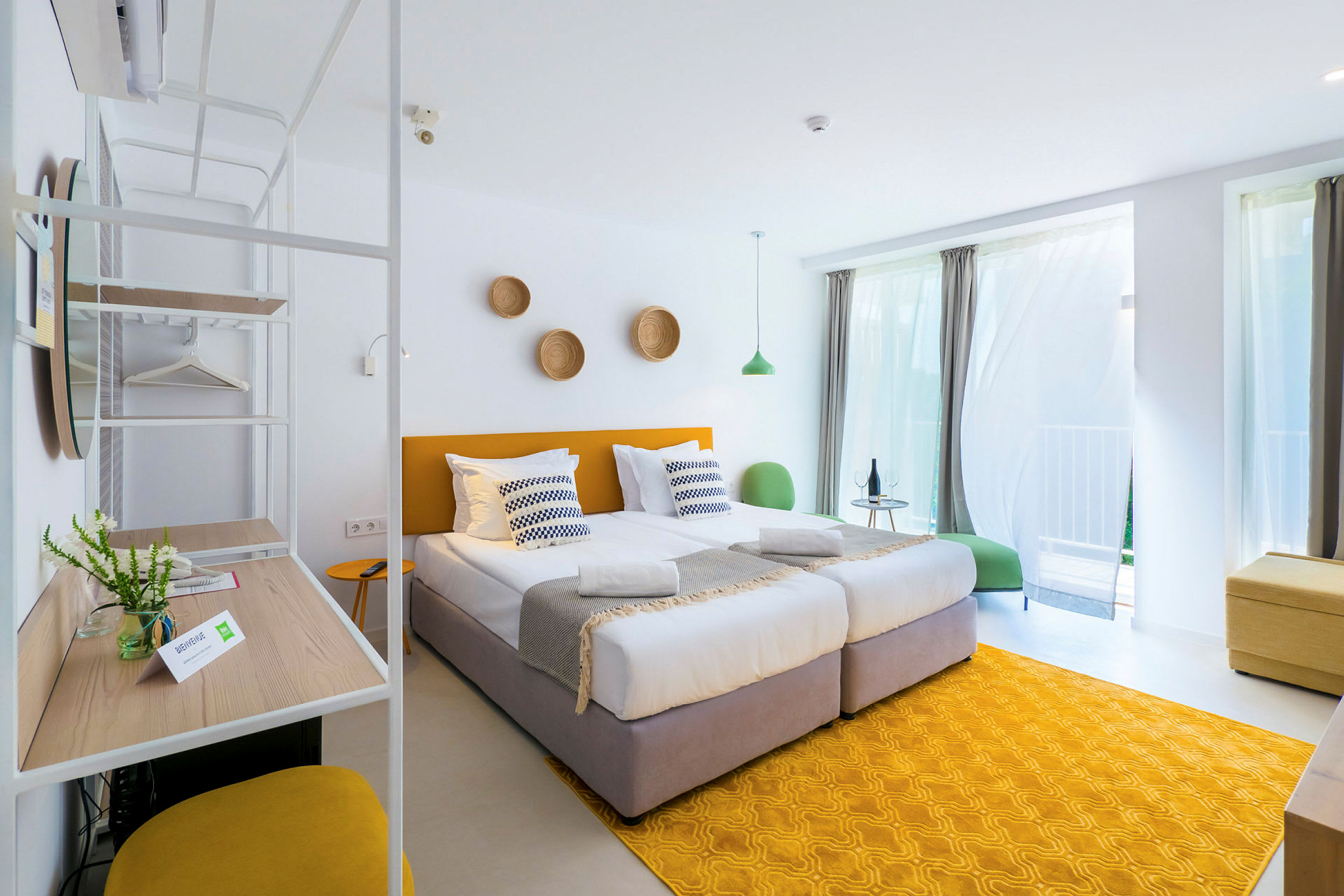 Ett ljust och modernt hotellrum med två enkelsängar, gula detaljer och stora fönster med gardiner. (AI-genererad text)