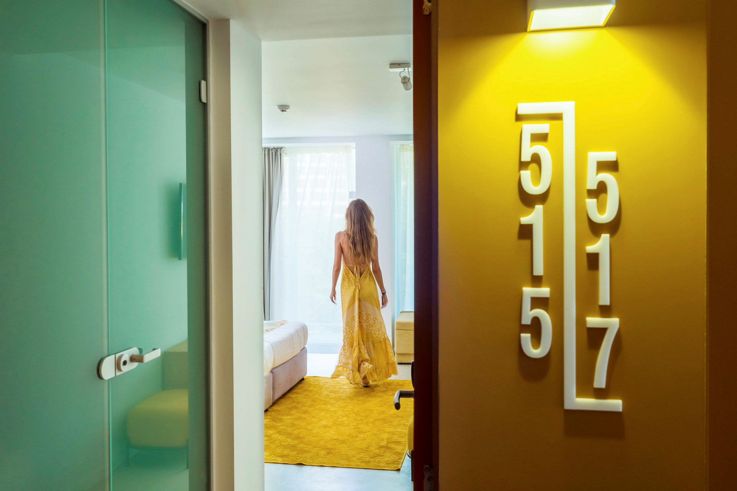 En person i en gul klänning som går in i ett hotellrum med ljus inredning. (AI-genererad text)