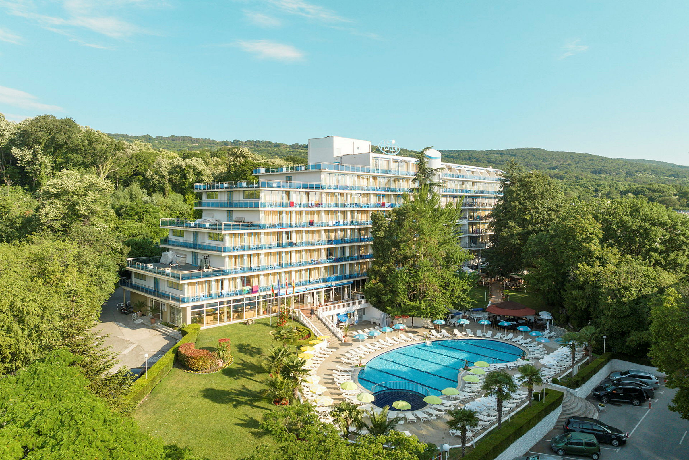 Bild av ett hotell med flera våningar och balkonger, omgiven av frodig grönska. En pool med solstolar ligger i förgrunden. (AI-genererad text)