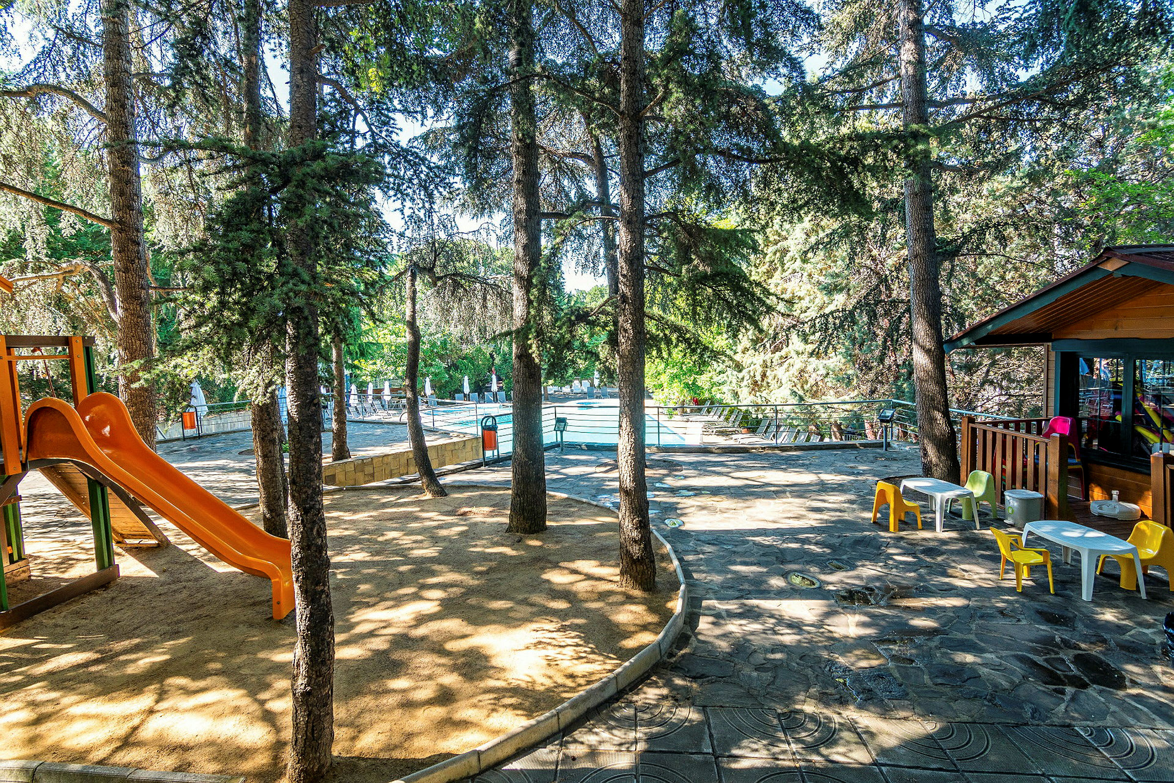 En lekplats med en orange rutschkana under trädens skugga nära en pool. (AI-genererad text)