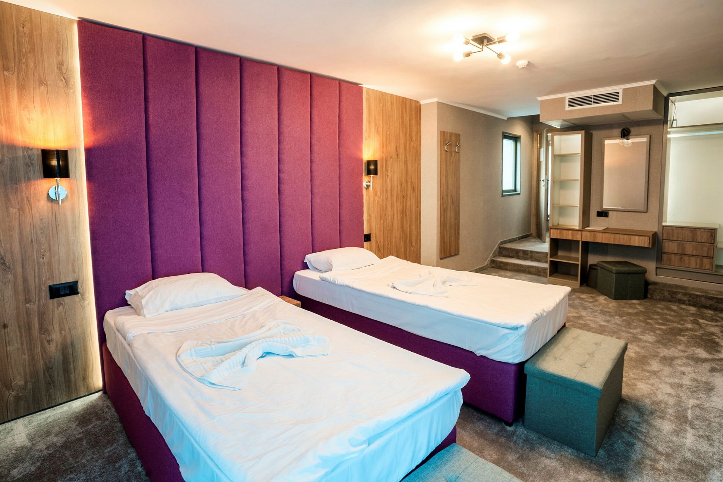 Två enkelsängar med vita lakan i ett hotellrum med lila sänggavel, träpaneler och en arbetsyta med spegel. (AI-genererad text)