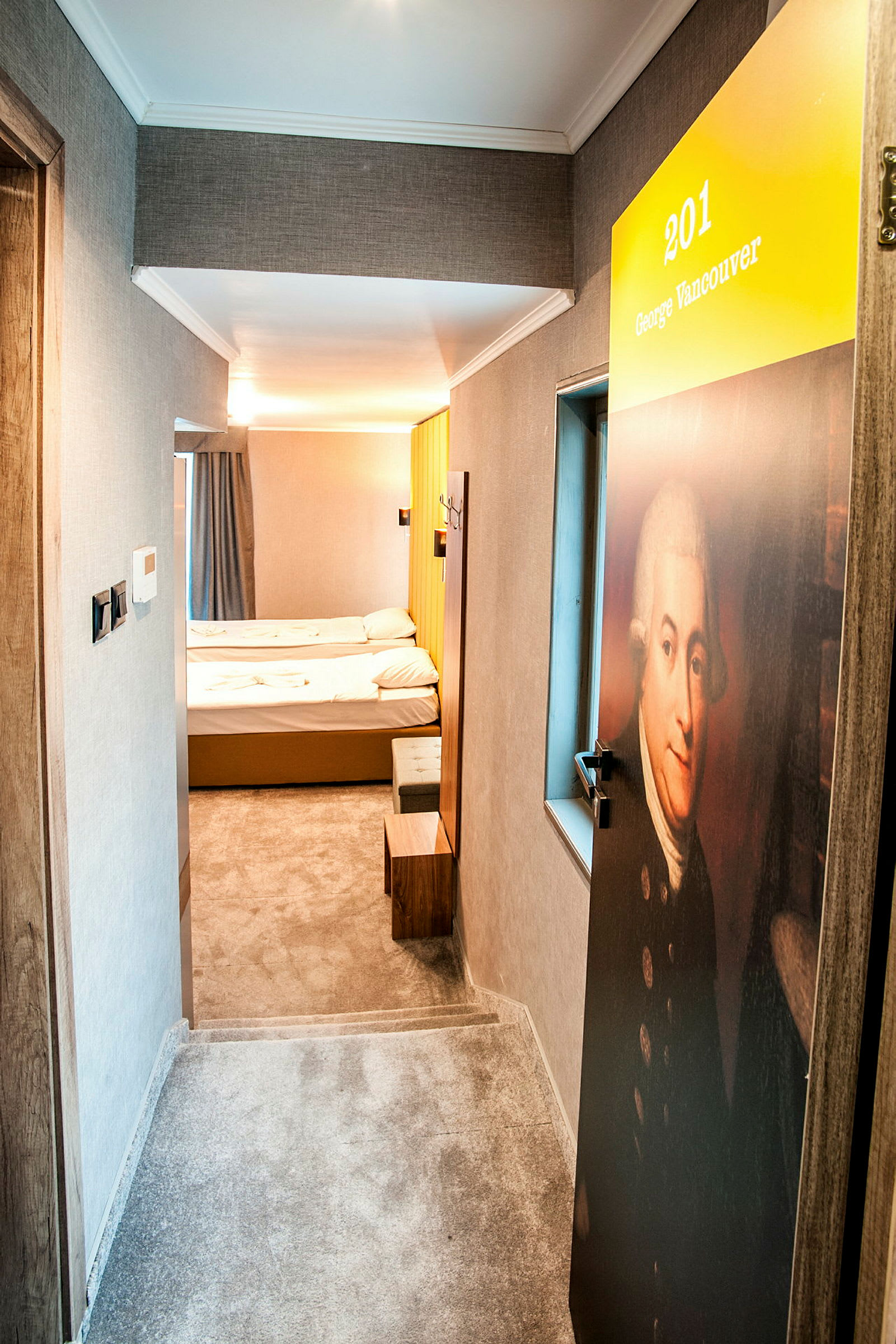En hotellkorridor med en dörr märkt med nummer 201 och namnet George Vancouver. Innanför syns en enkel hotellrumsmiljö med två sängar. (AI-genererad text)