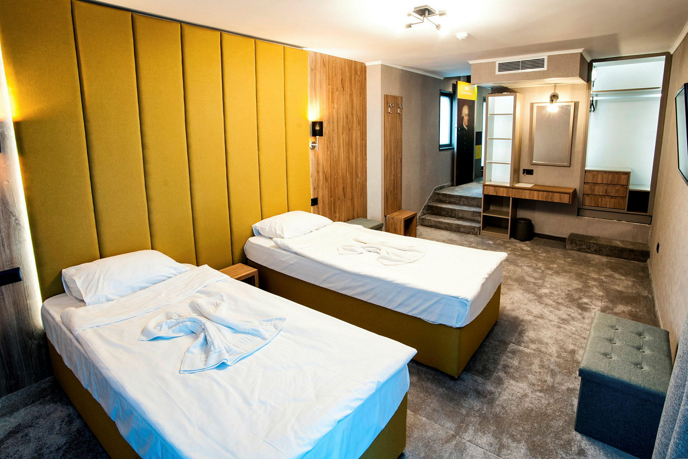 Ett hotellrum med två enkelsängar med gul klädsel och vita sängkläder. Rummet har en modern inredning med en gul väggpanel, ett skrivbord, sittmöbler och en liten förvaringshylla. (AI-genererad text)