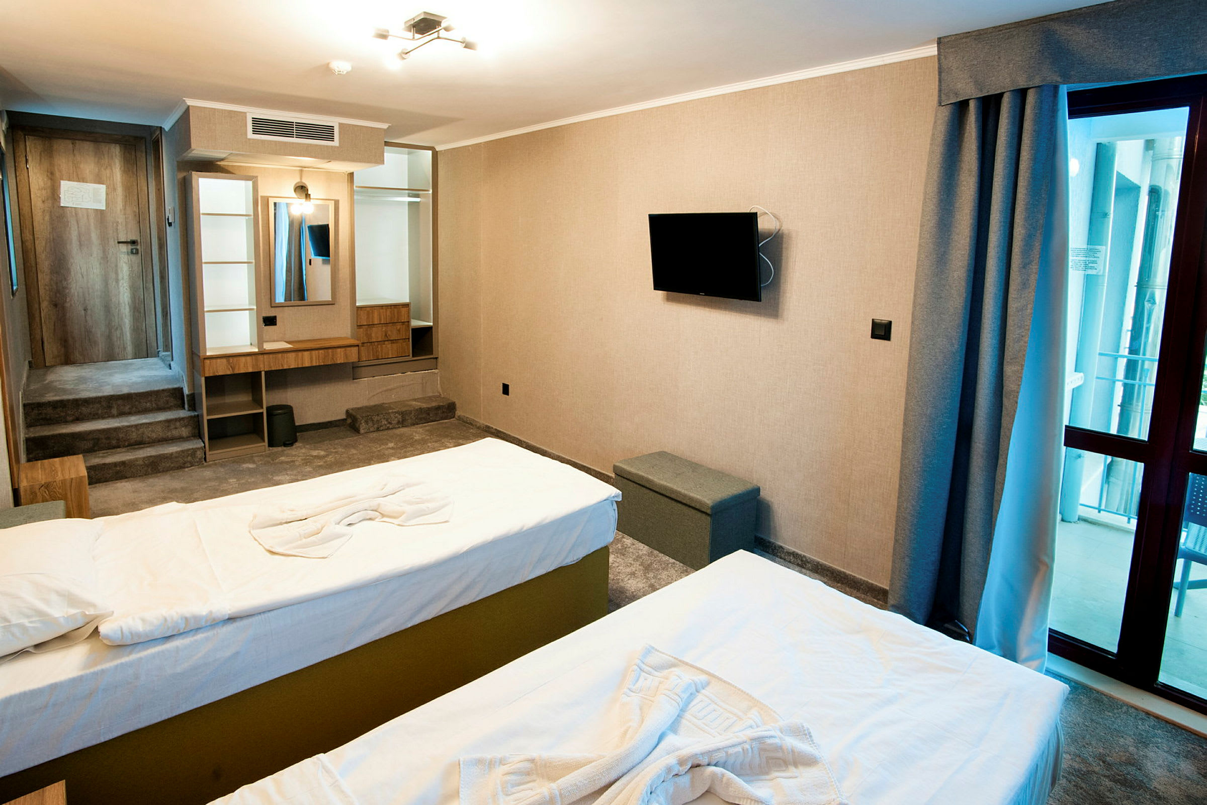 Ett hotellrum med två sängar, en väggmonterad TV, spegel med skrivbord och en glasdörr mot balkong. (AI-genererad text)