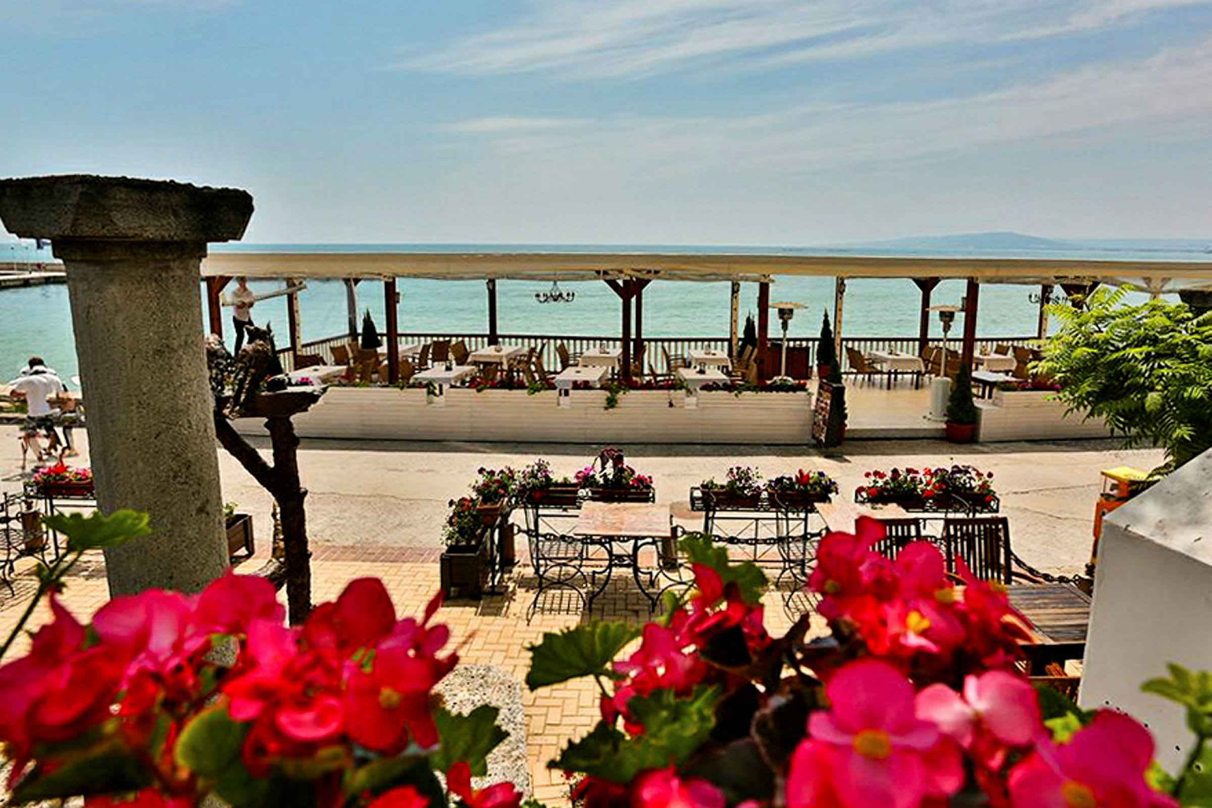 En strandnära terrass med bord och stolar som omges av blommor och utsikt över havet. (AI-genererad text)