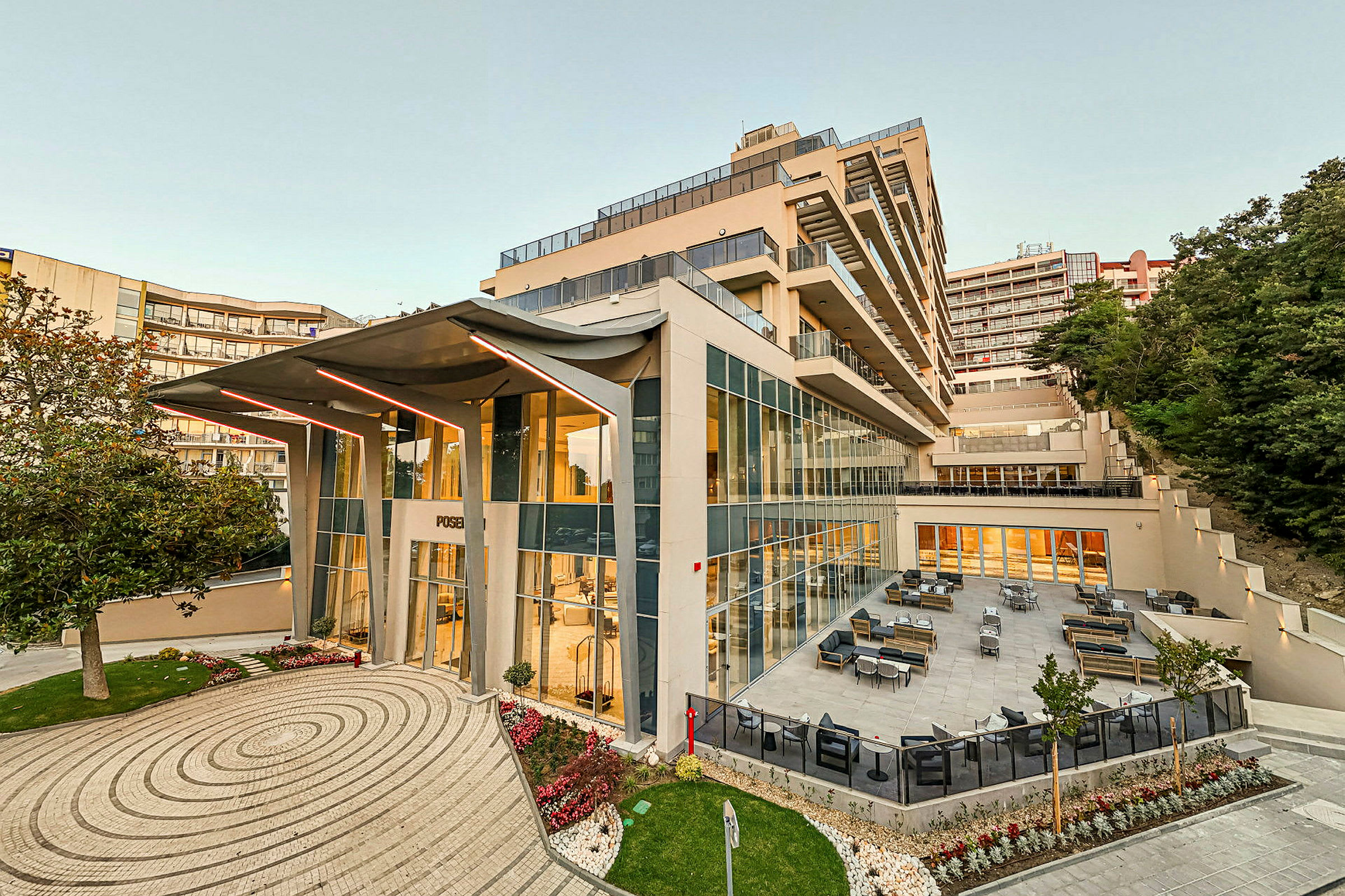 En modern hotellbyggnad med glasfasad och en utomhusterrass med sittmöbler, omgiven av växtlighet. (AI-genererad text)