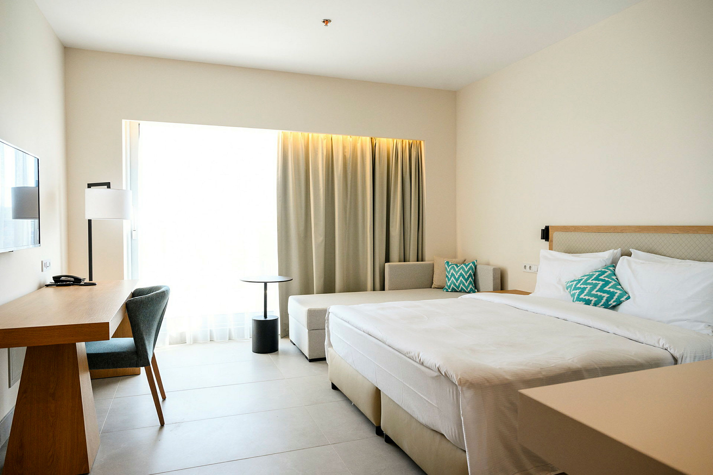 Ett modernt hotellrum med en dubbelsäng, arbetsbord, stolar och stora fönster med ljusa gardiner. (AI-genererad text)