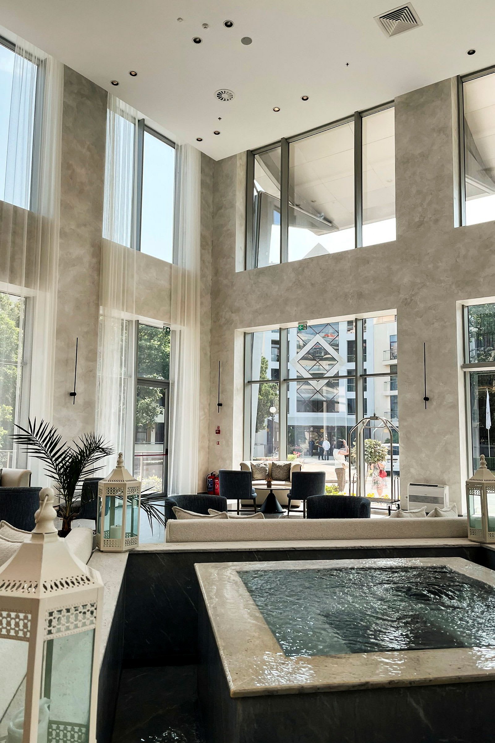 Ett modernt hotelllobby med stora fönster som släpper in dagsljus. Det finns bekväma sittplatser och ett vattenelement i förgrunden. (AI-genererad text)