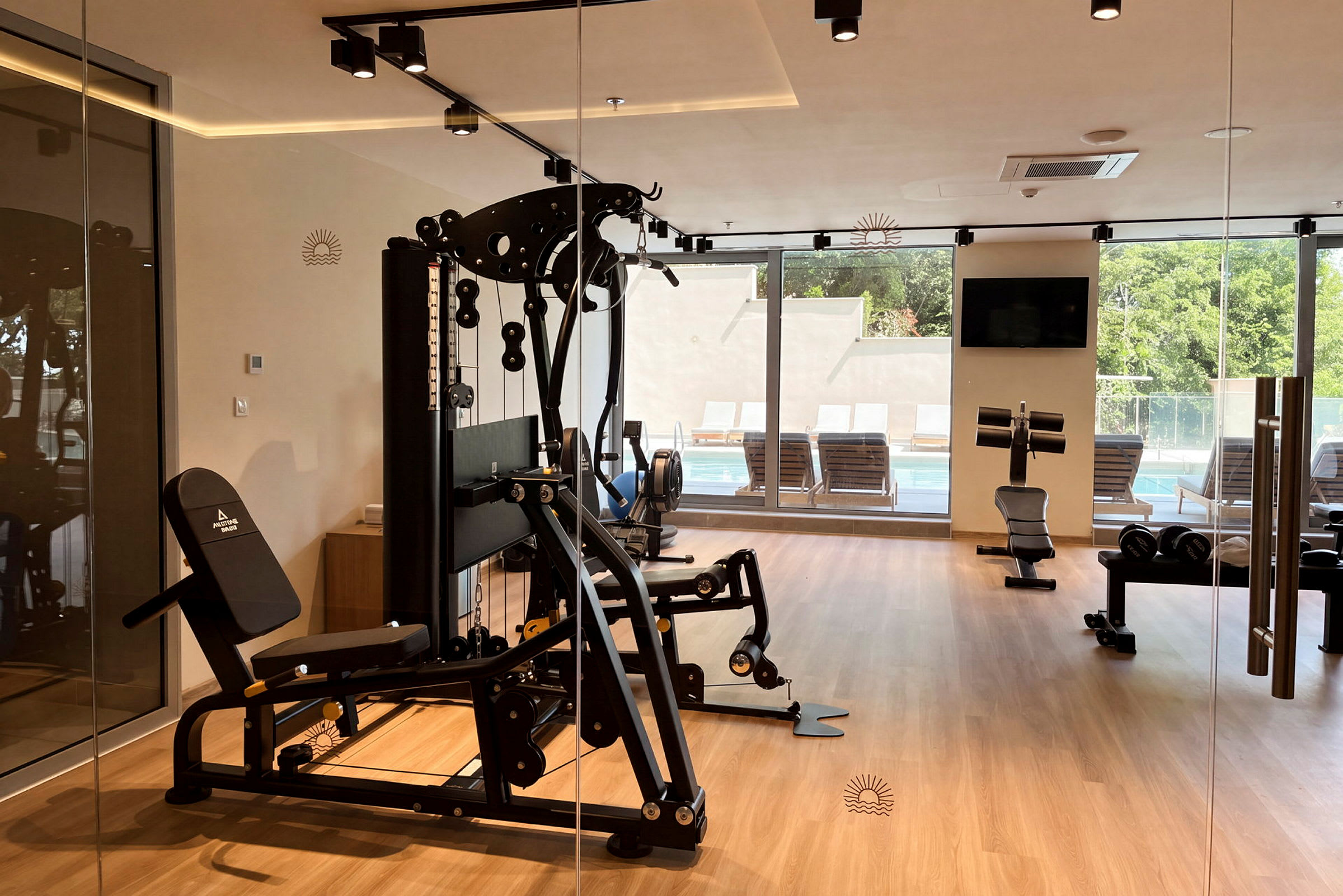 Ett gym med träningsmaskiner och fönster som vetter mot en pool och grönskande omgivning. (AI-genererad text)