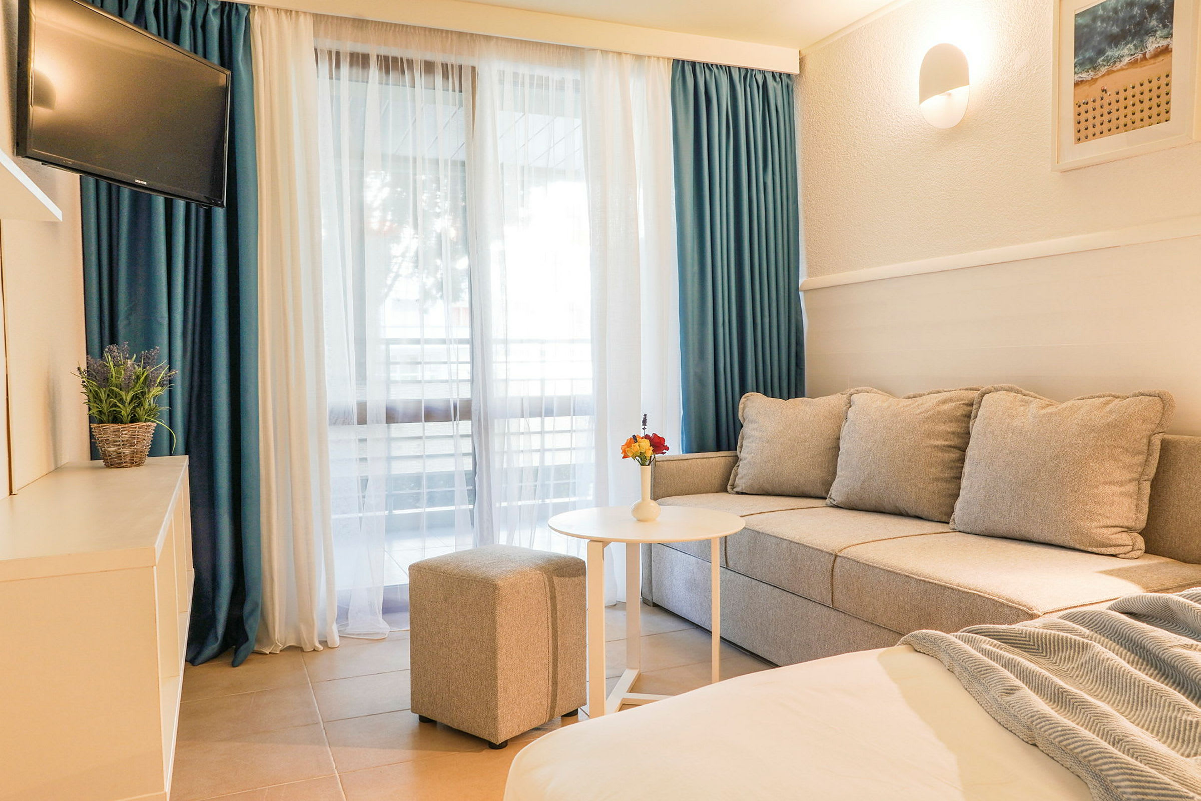 Ett hotellrum med soffa, bord, ottoman och TV, samt ett stort fönster med gardiner. (AI-genererad text)