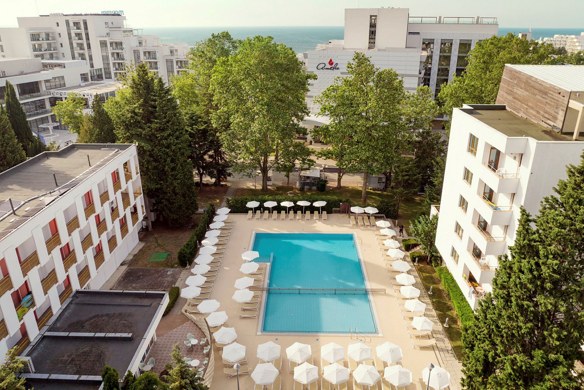 En pool omgiven av solstolar och parasoller, med hotellbyggnader och träd i bakgrunden. (AI-genererad text)