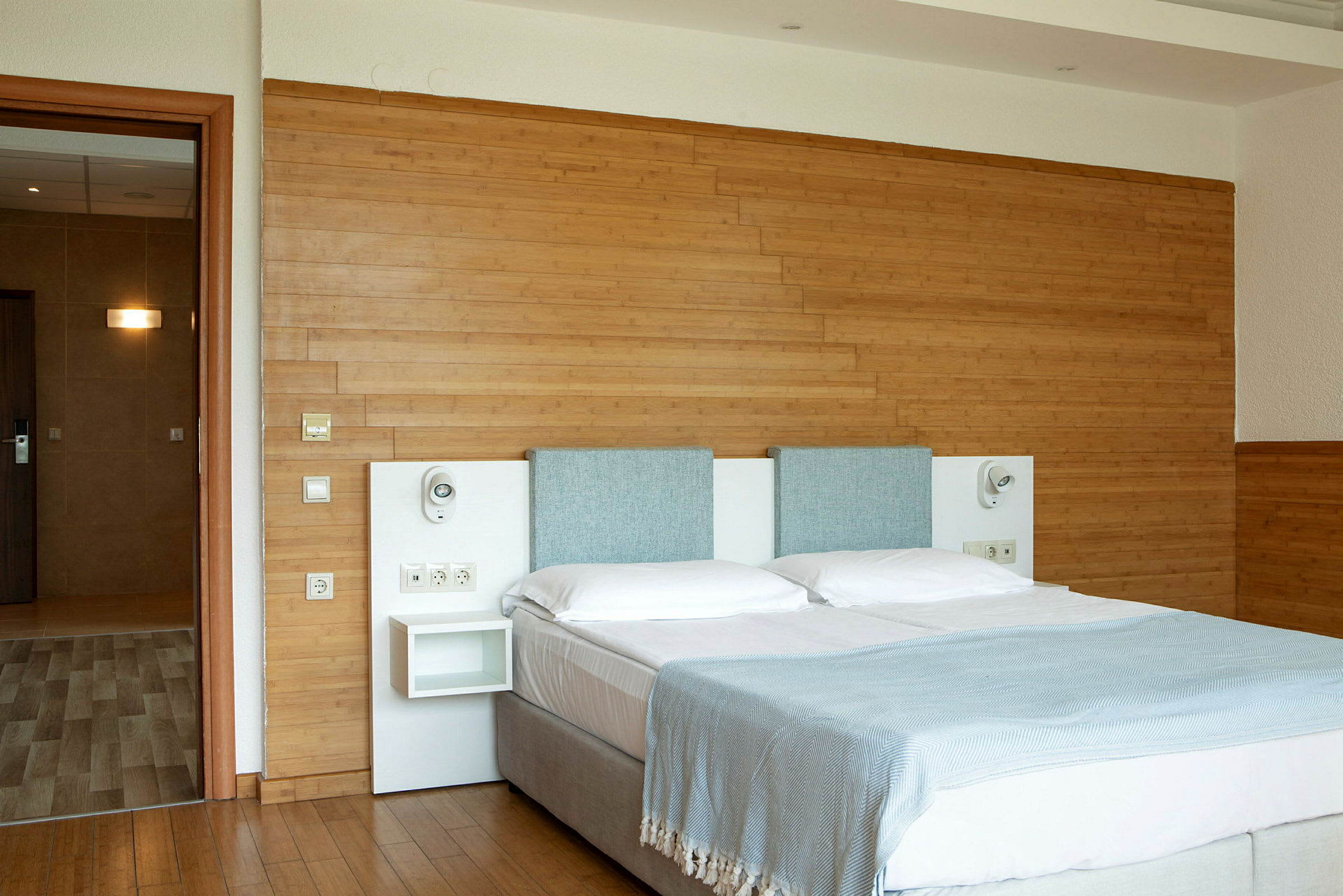 Ett hotellrum med en dubbelsäng klädd i vita lakan och ljusblå överkast. Bakom sängen finns en träpanel på väggen. (AI-genererad text)