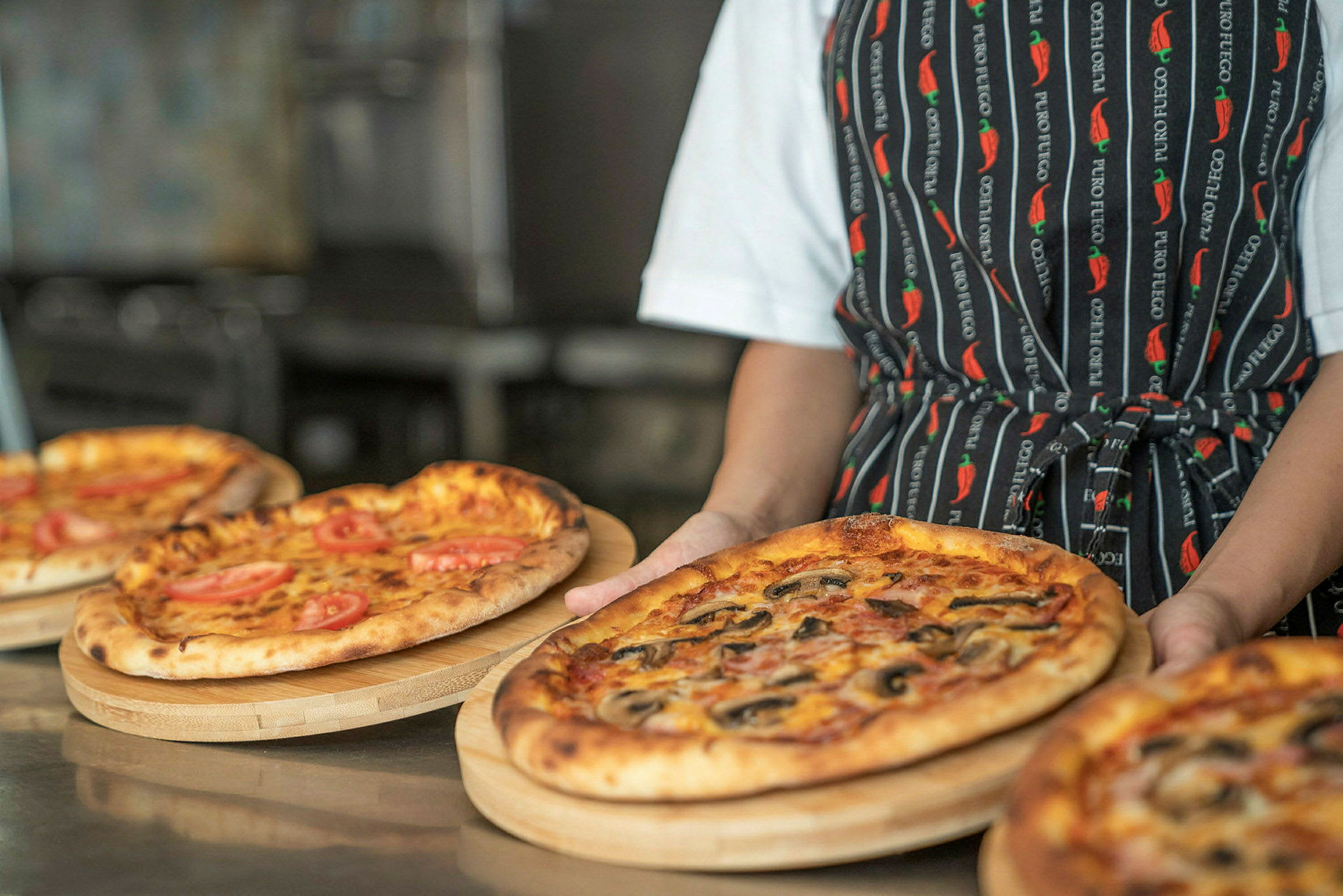 En person håller i en träbricka med en pizza, omgiven av andra pizzor. (AI-genererad text)
