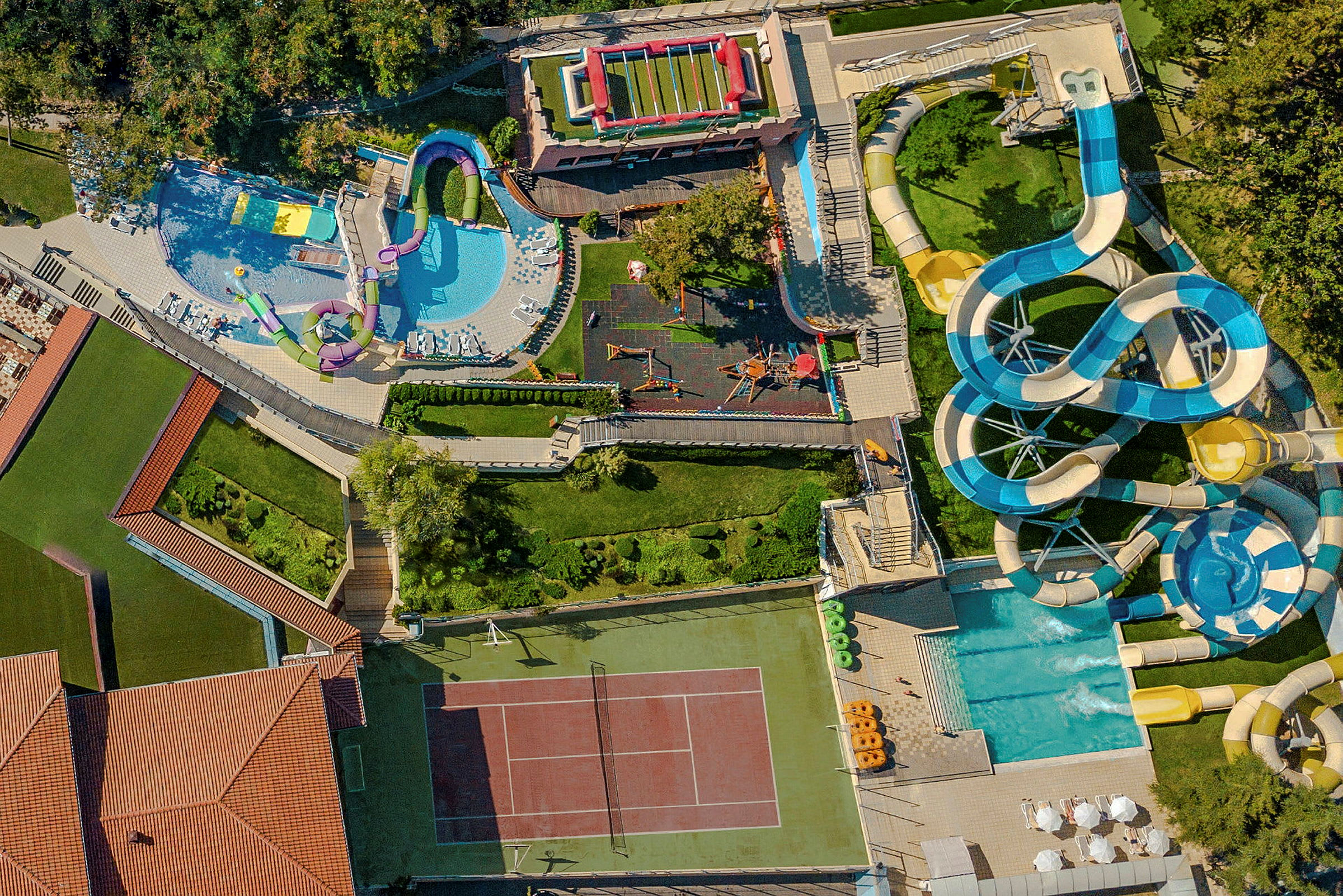 Flygfoto av en anläggning med en tennisbana, pooler, vattenrutschbanor och lekplatser, omgivna av grönområden. (AI-genererad text)