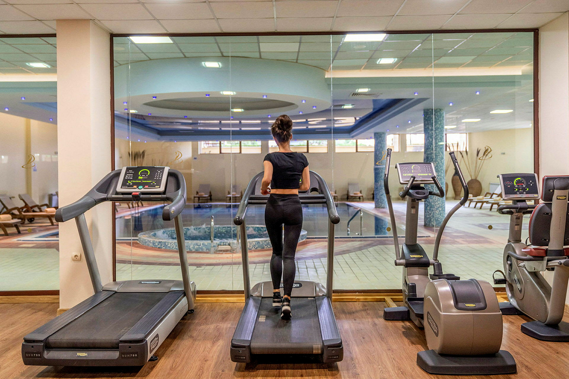 Kvinna tränar på ett löpband i ett gym med utsikt mot en inomhuspool. (AI-genererad text)