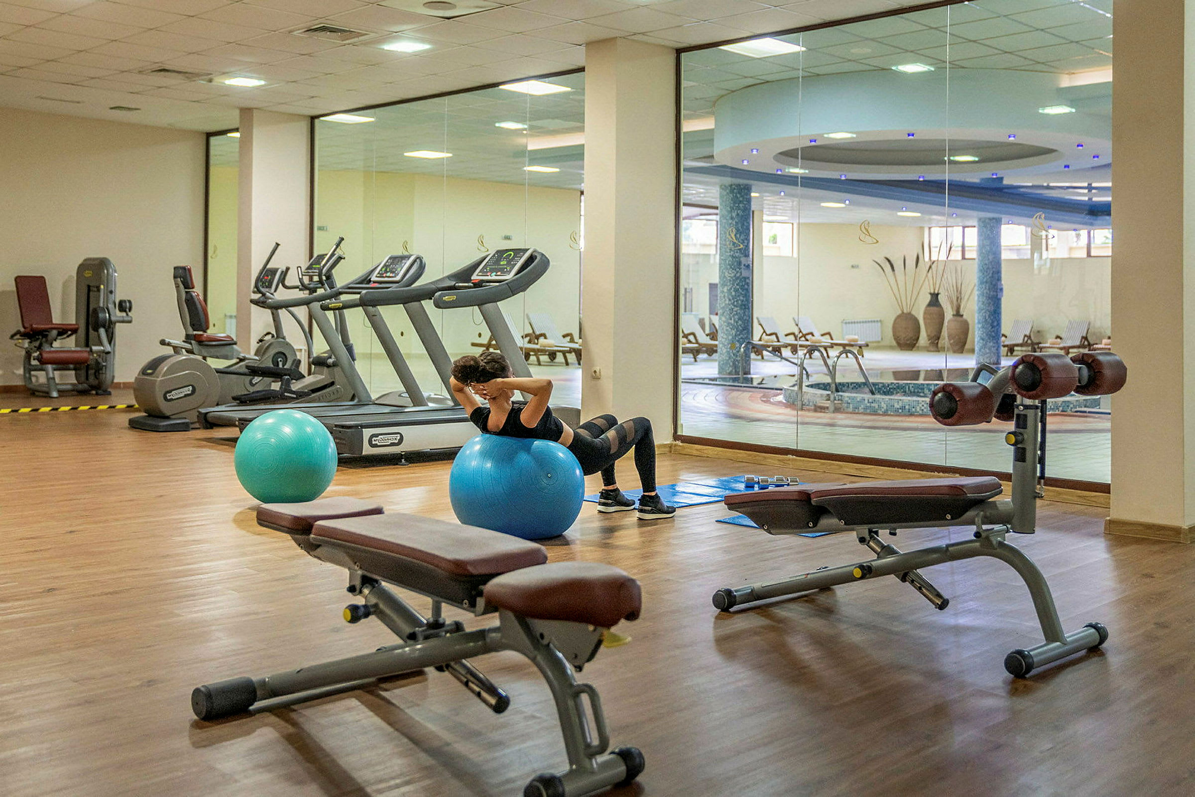 En person tränar på en blå träningboll i ett gym med träningsutrustning och utsikt mot en inomhuspool. (AI-genererad text)