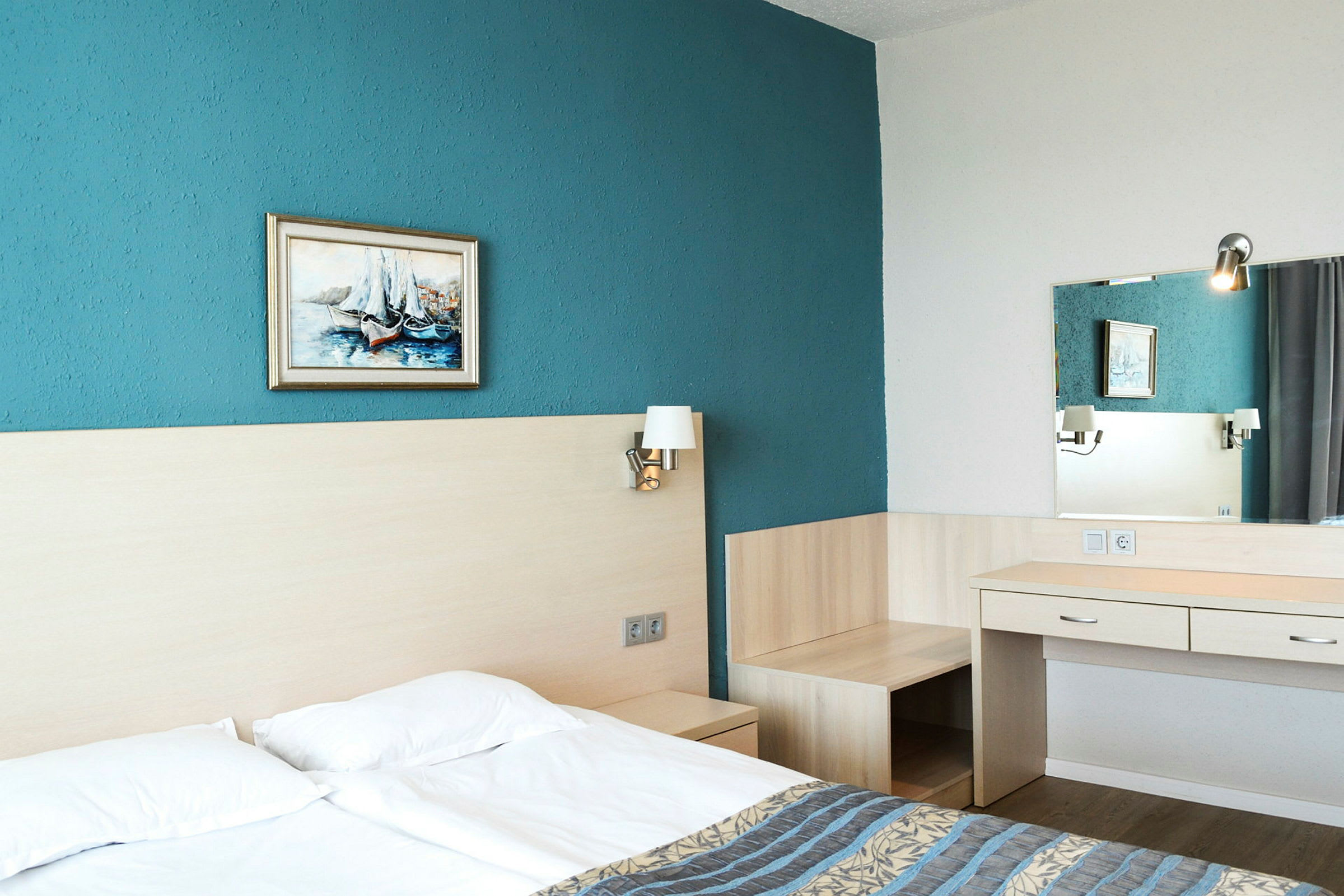 Hotellrum med dubbelsäng, blå vägg, tavla, sänglampor och skrivbord med spegel. (AI-genererad text)