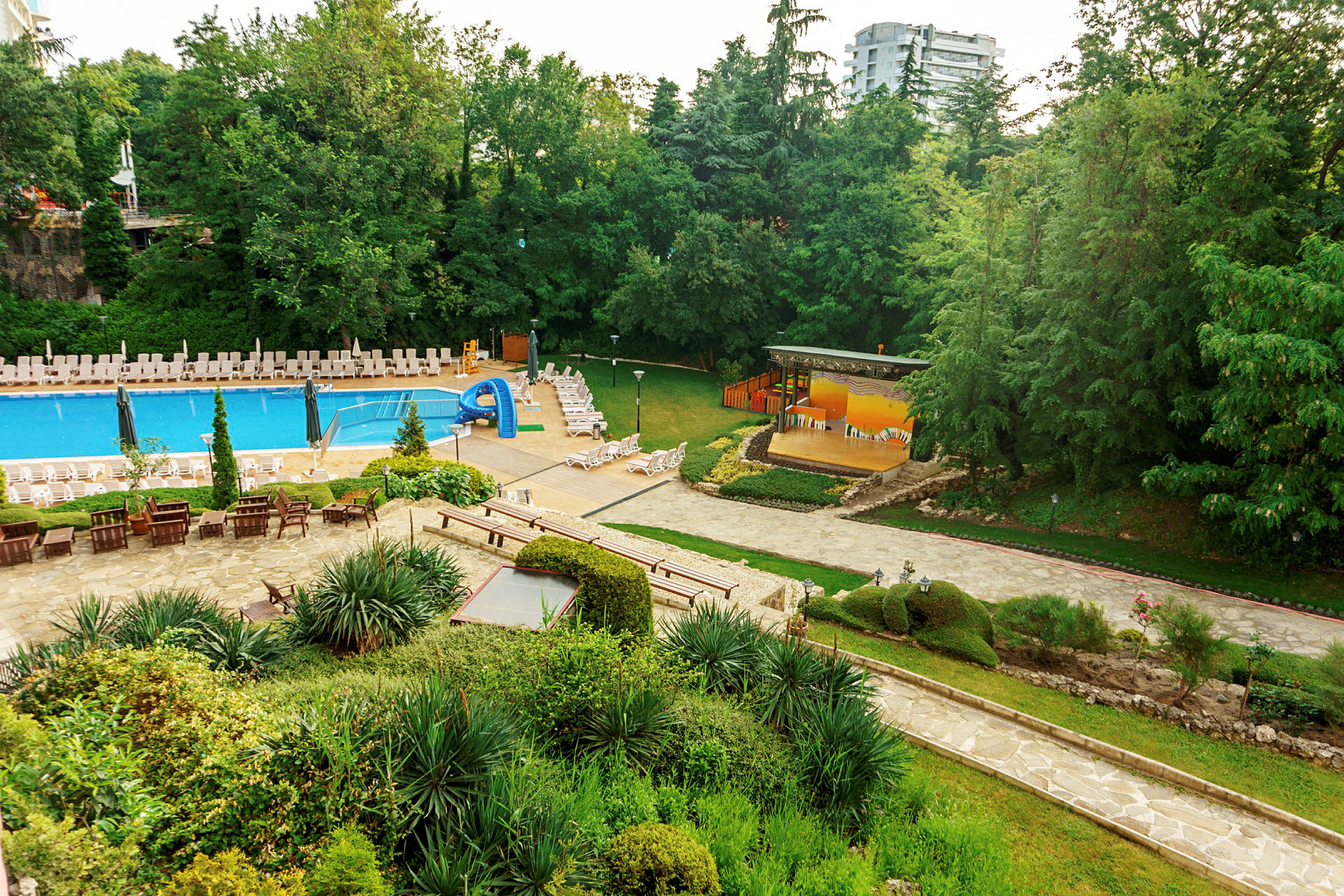 Utomhusområde med pool, solstolar, gröna växter och ett scenområde. (AI-genererad text)
