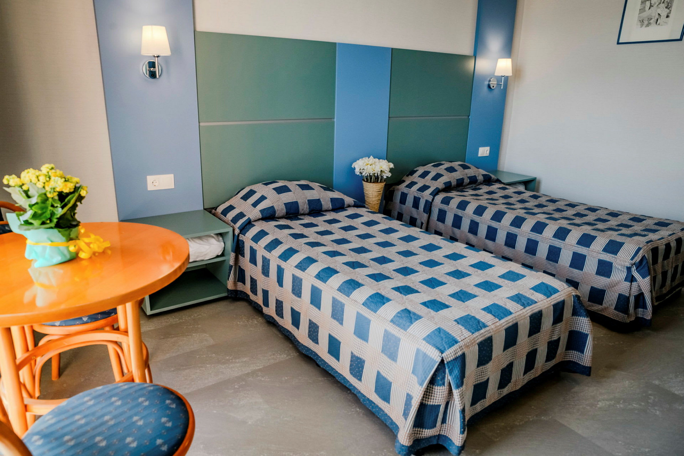 Ett hotellrum med två enkelsängar med blåmönstrade överkast, ett runt träbord med stolar samt dekorationer på sängbordet. (AI-genererad text)