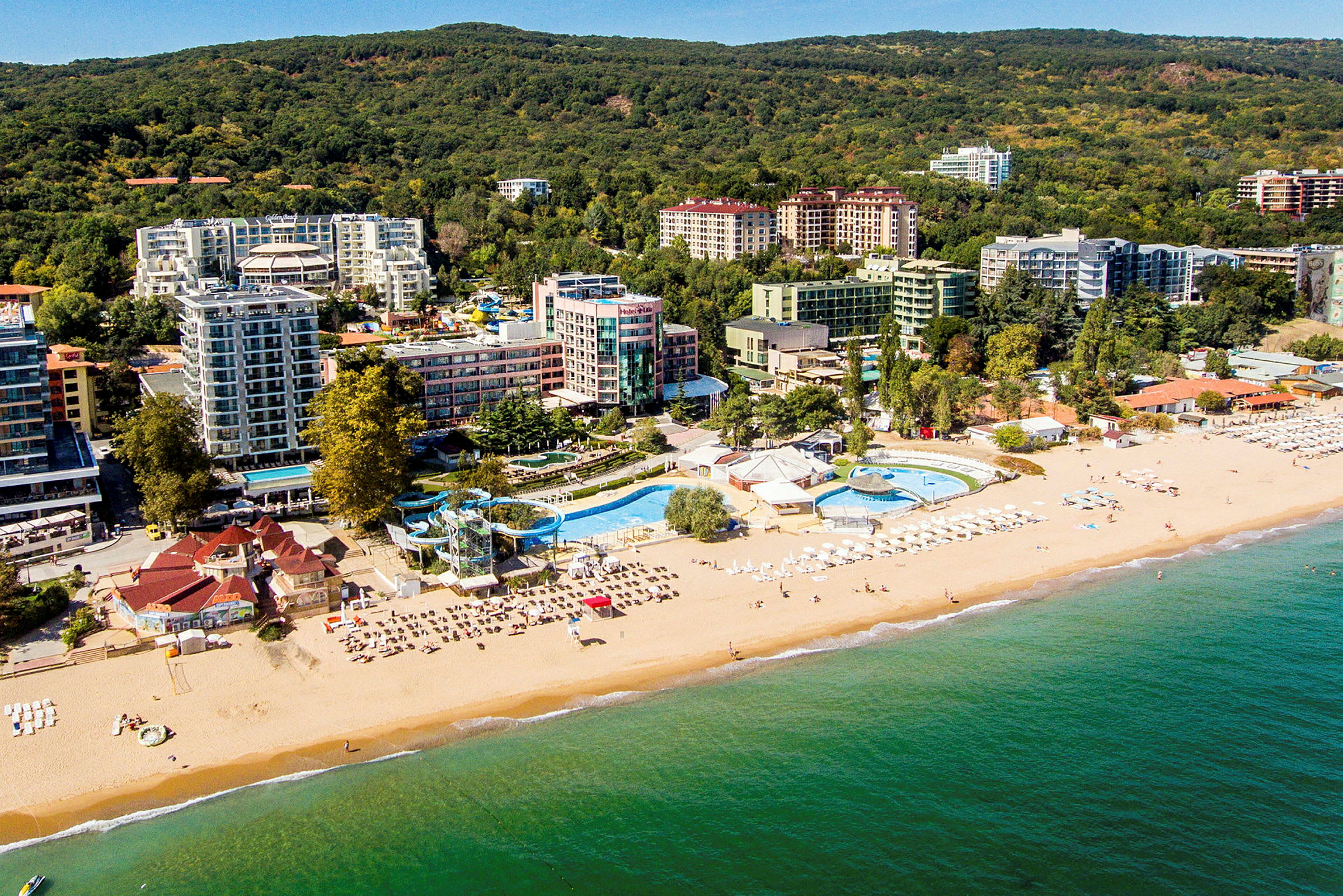 Flygbild av en strand med solstolar och parasoller, en poolanläggning och hotell omgivna av grönområden. (AI-genererad text)