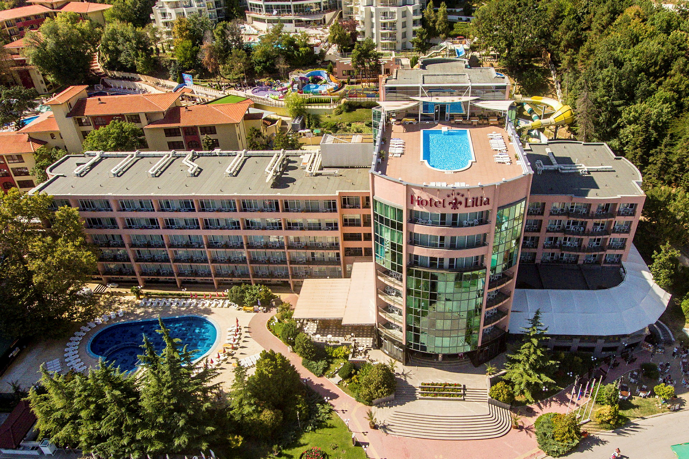 En hotellbyggnad med pool på taket och en pool på markplan, omgiven av grönska. (AI-genererad text)