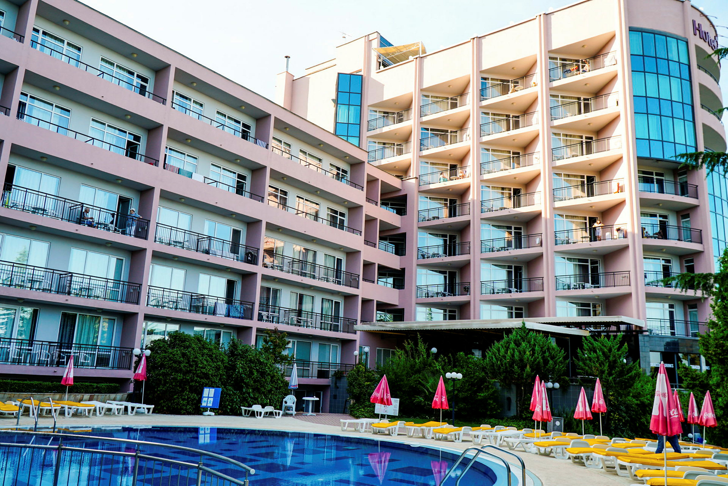Ett större hotell med flera våningar och balkonger med utsikt över en pool samt solstolar och parasoller. (AI-genererad text)