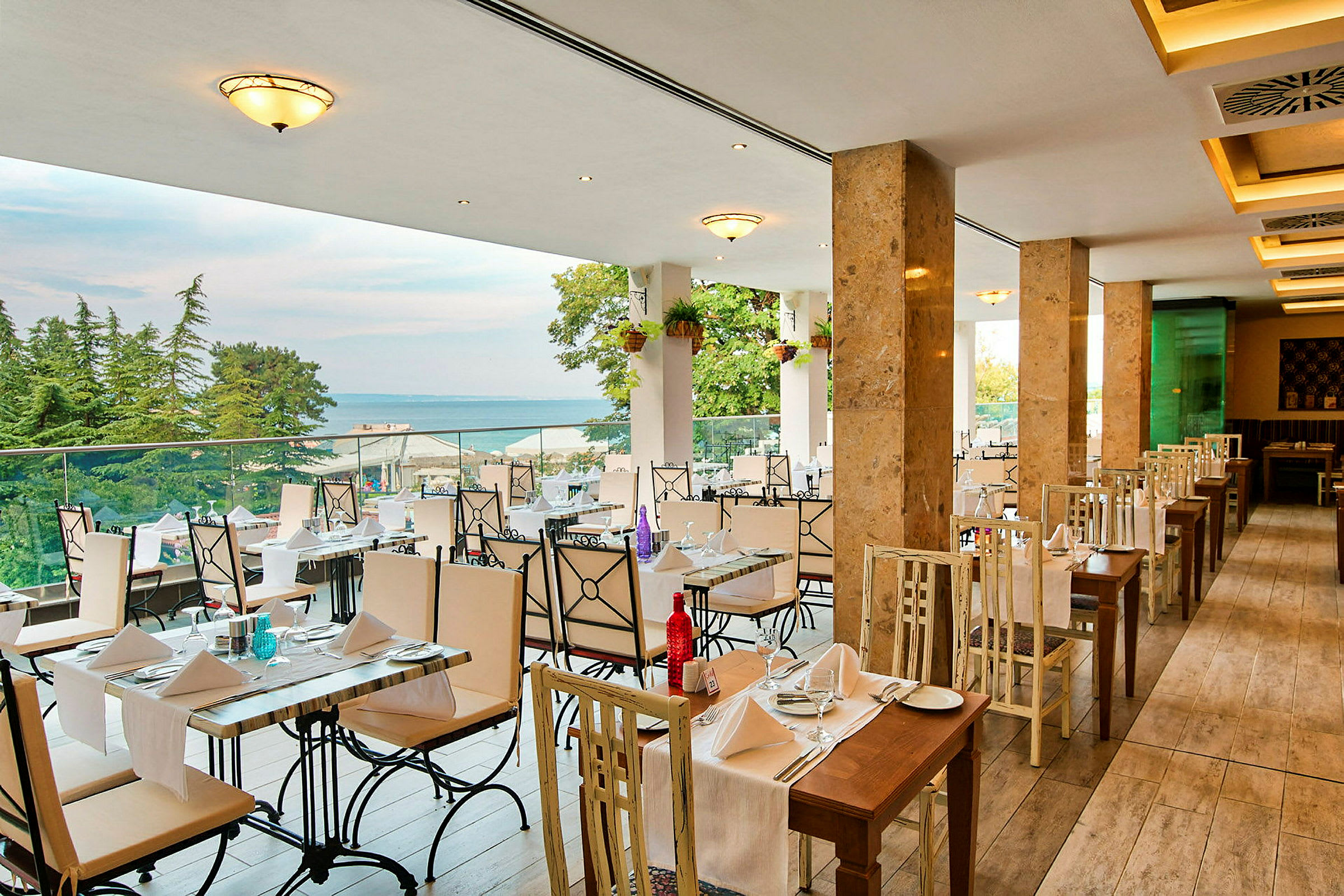 En restaurang med bord och stolar i ljusa färger, utsikt mot havet och omgiven av träd. (AI-genererad text)