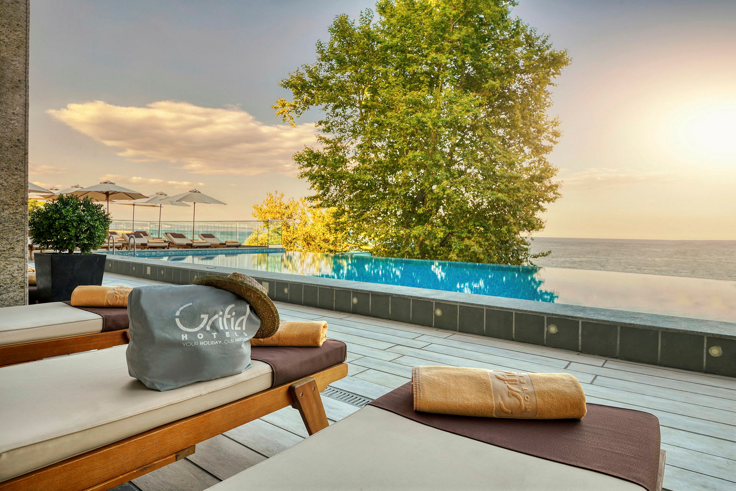 Solstolar med handduk och hatt vid en infinitypool, träd och hav i bakgrunden. (AI-genererad text)