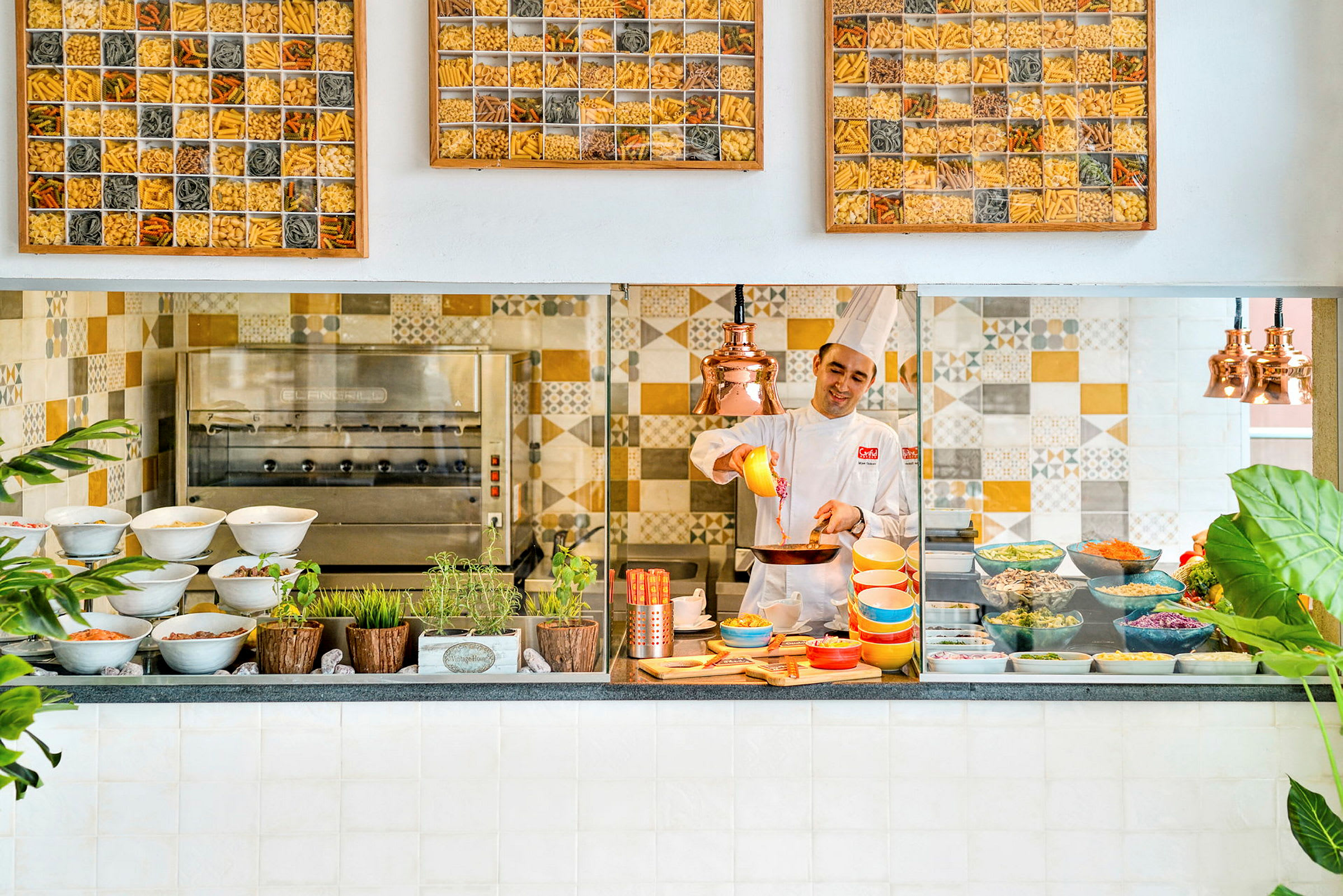 Bild på en köksmiljö med en professionell kock som arbetar med olika färgglada skålar och ingredienser. Köket har dekorativa kakel och växter. (AI-genererad text)