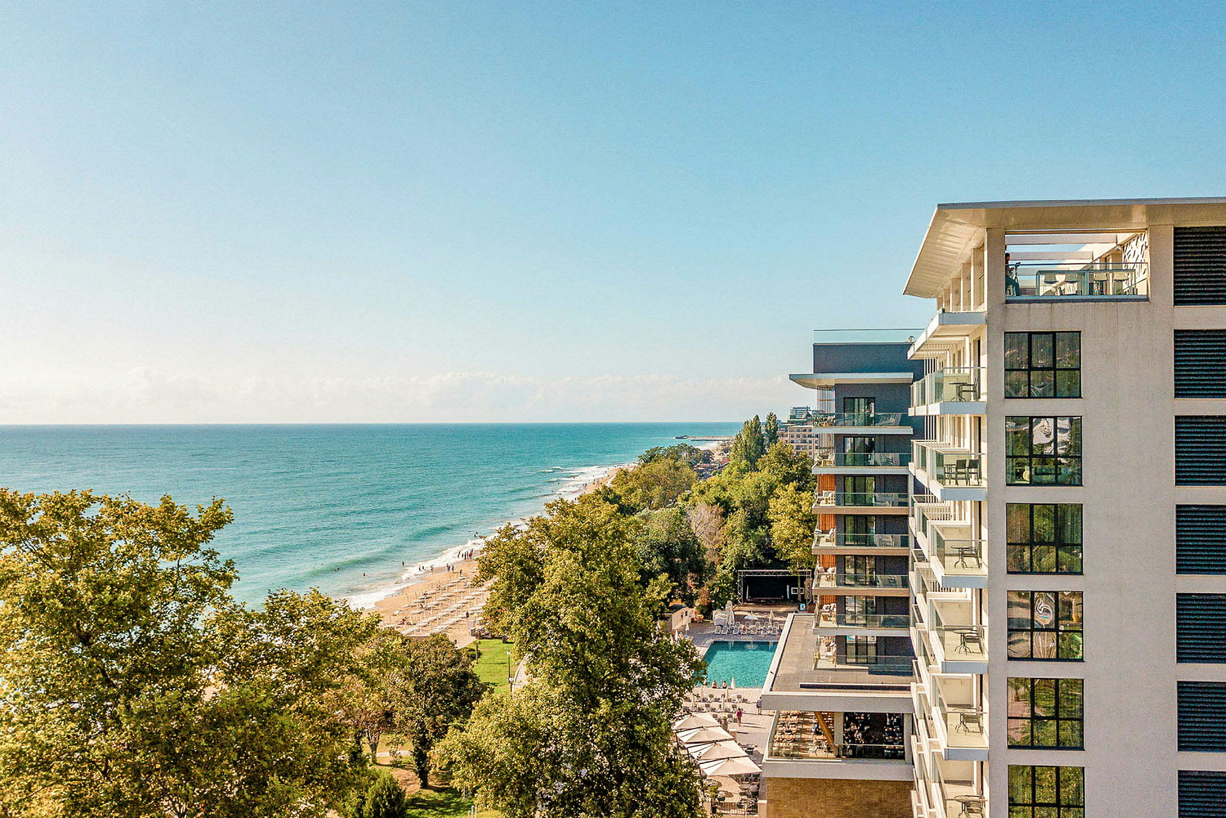 En vy från höjd över havskusten med ett hotell på höger sida och stranden omgiven av träd. (AI-genererad text)