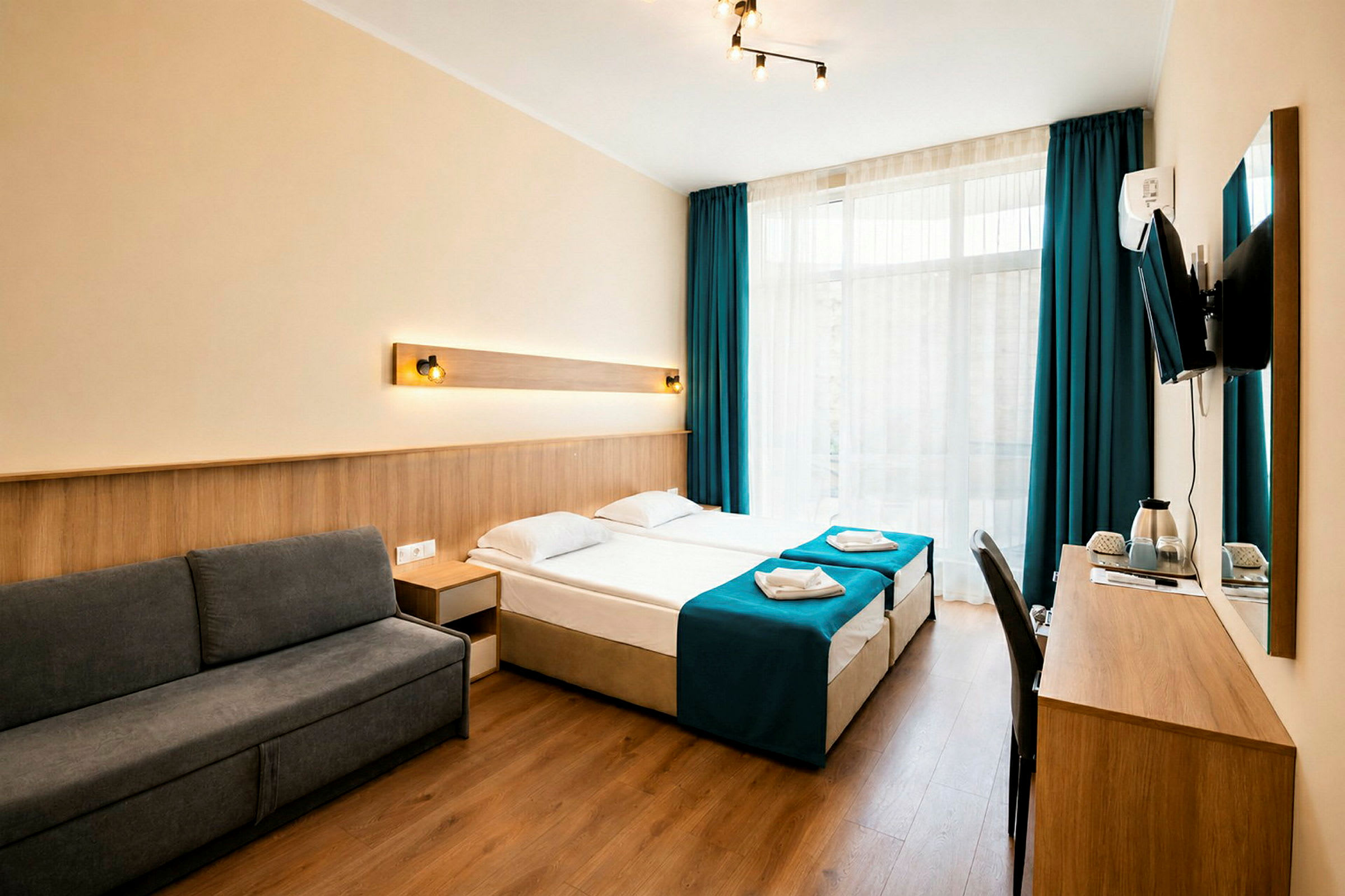 Ett hotellrum med en modern inredning, mörkgrå soffa, två enkelsängar med blå överkast och ett skrivbord med en väggmonterad TV. (AI-genererad text)