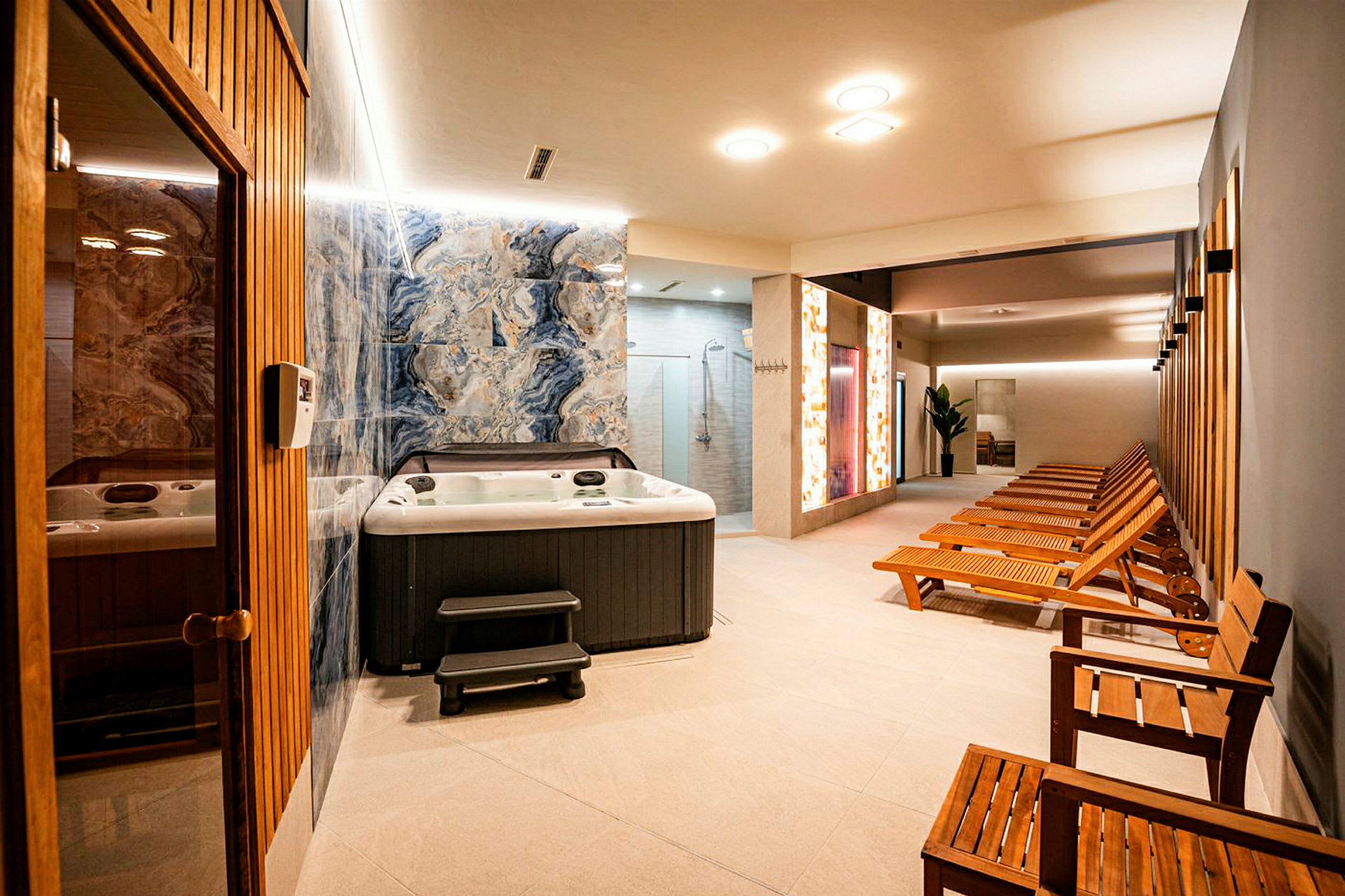 En spa-avdelning med en jacuzzi, trästolar och solstolar, samt en dörr till en bastu. (AI-genererad text)
