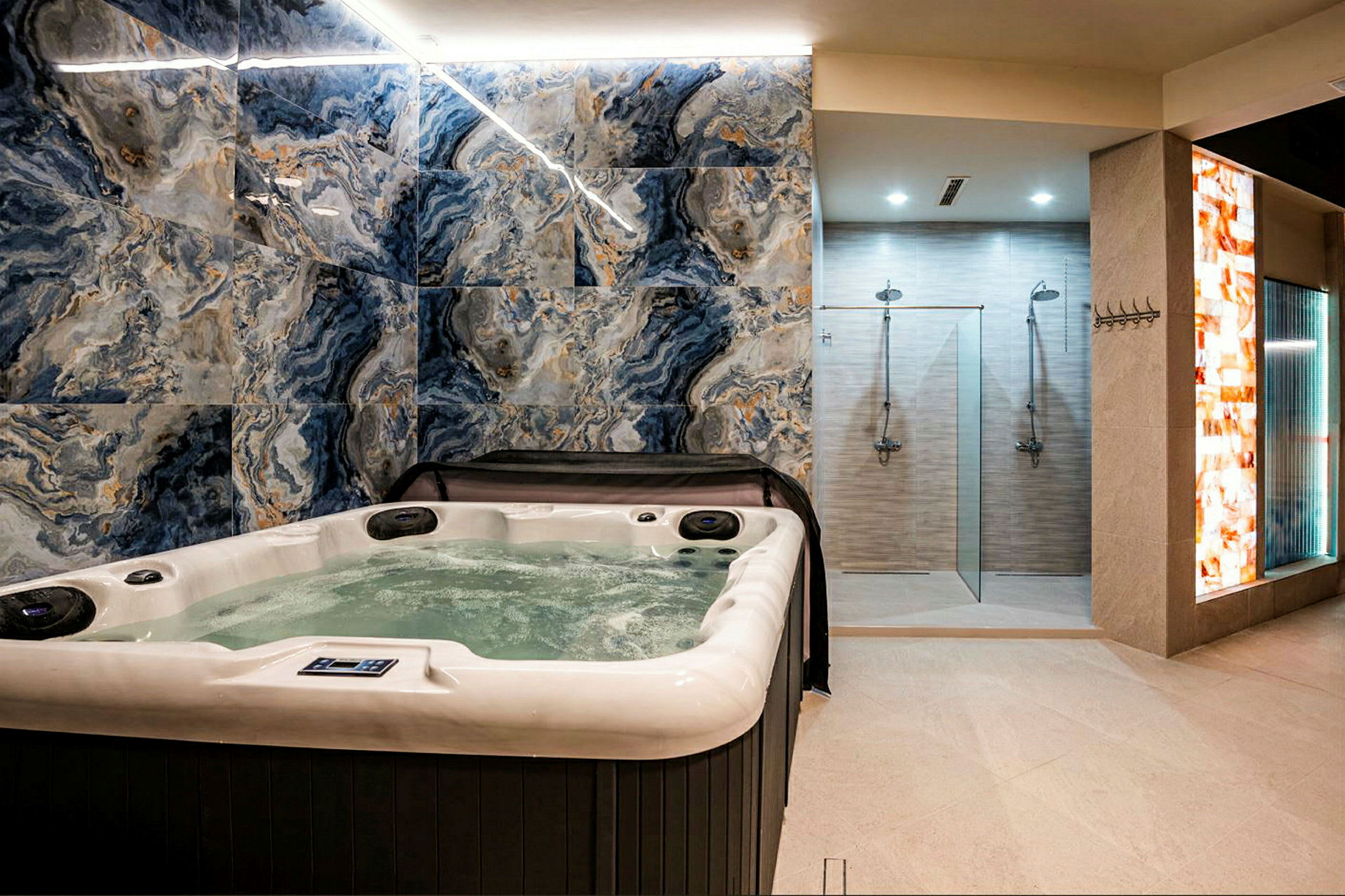 Ett spaområde med en jacuzzi i förgrunden och duschar i bakgrunden, omgiven av moderna väggar med marmormönster. (AI-genererad text)