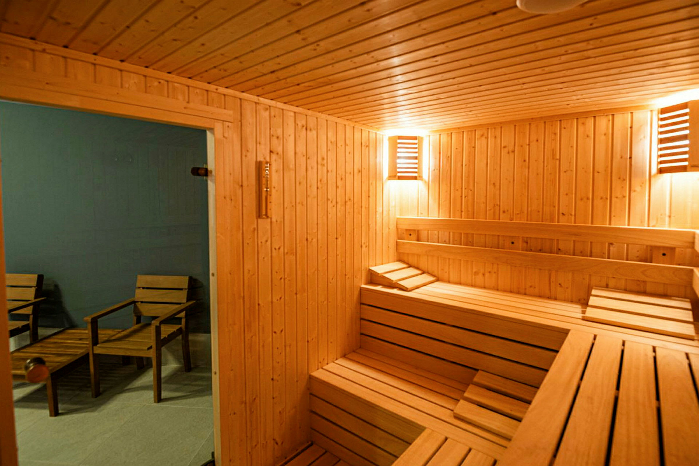 Ett rum inrett som en traditionell bastu med träpanel och sittplatser i trä. Utanför bastun syns stolar och ett bord. (AI-genererad text)