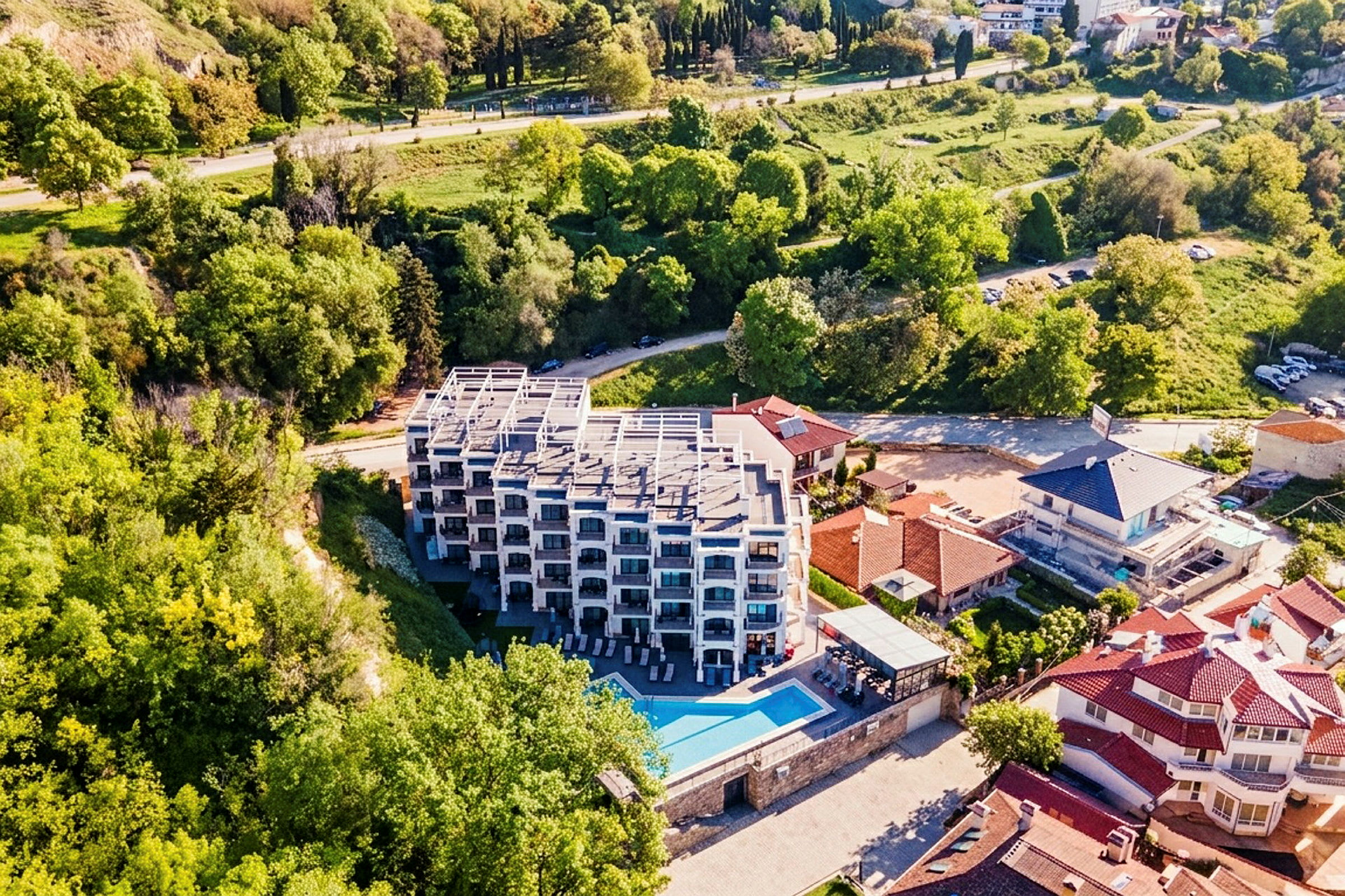 Ett hotell i flera våningar med balkonger och en pool framför, omgivet av gröna områden och andra hus. (AI-genererad text)