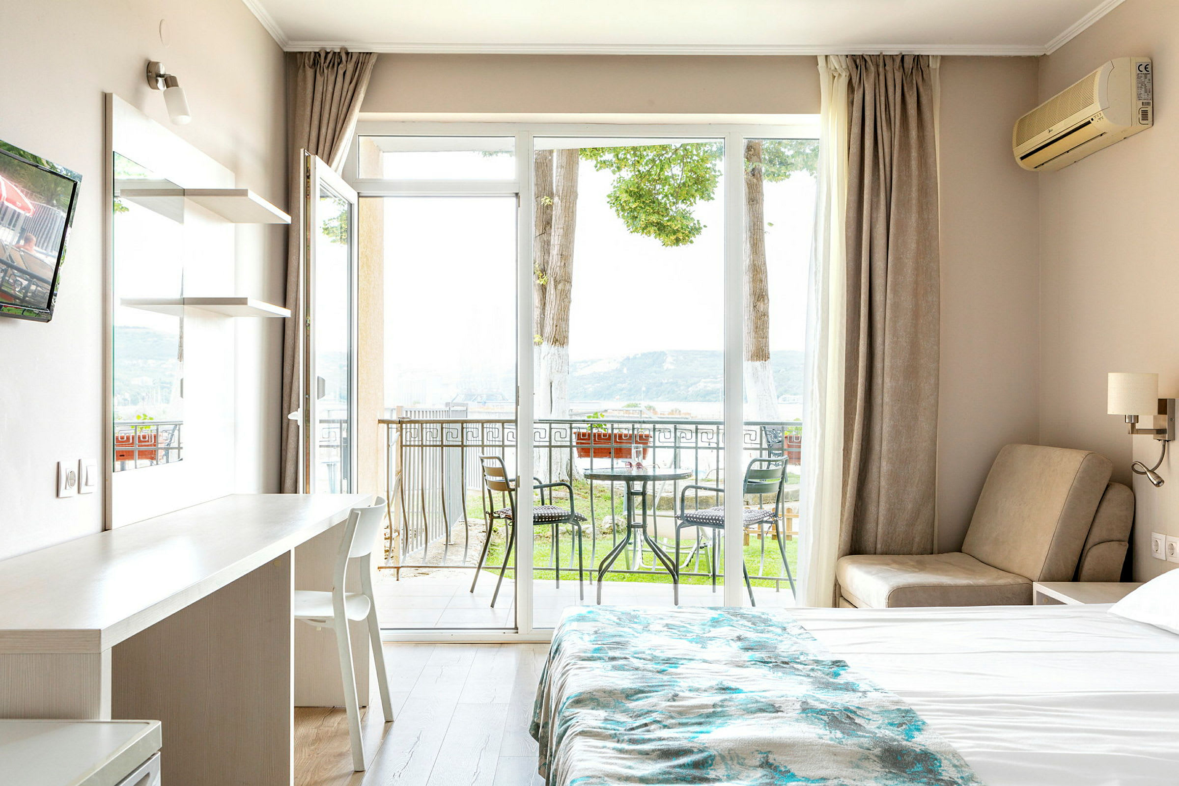 Ett hotellrum med en säng, ett bord och en stol, samt en balkong med utemöbler och utsikt mot naturen. (AI-genererad text)