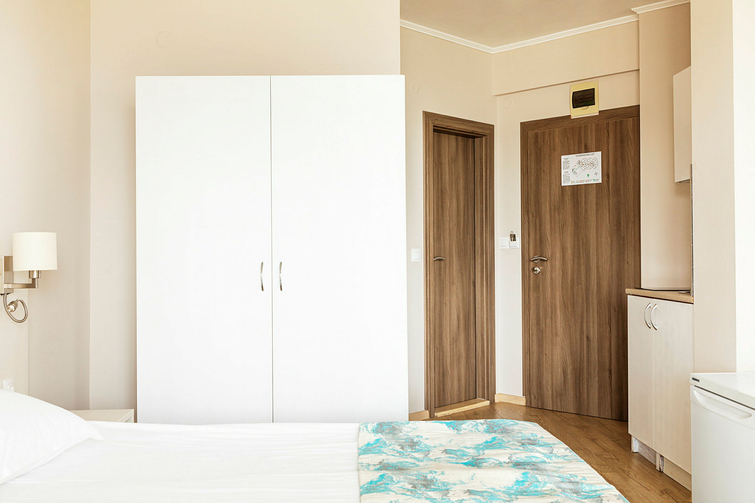 En hotellrumsmiljö med en vit garderob, trädörrar och del av en säng i förgrunden. (AI-genererad text)