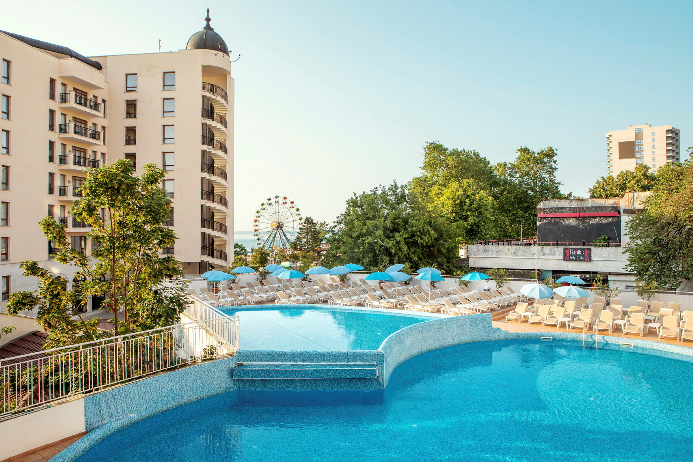 Hotellbyggnad med balkonger, en utomhuspool och solstolar med parasoller. Bakgrunden visar gröna träd och ett pariserhjul. (AI-genererad text)