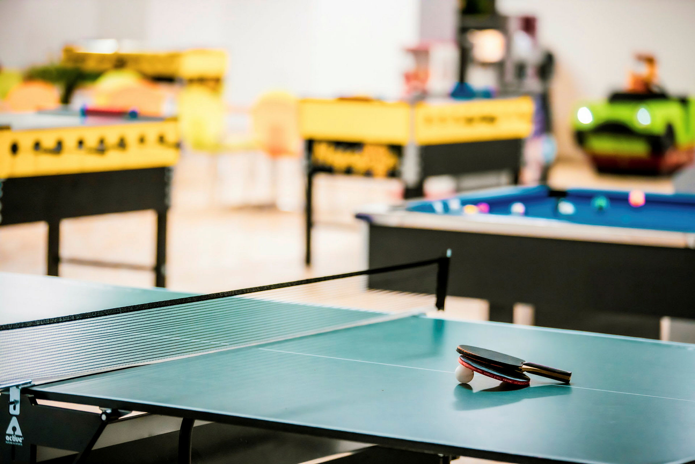 En pingisbord med racketar och boll, med biljard- och bordsfotbollsbord i bakgrunden. (AI-genererad text)
