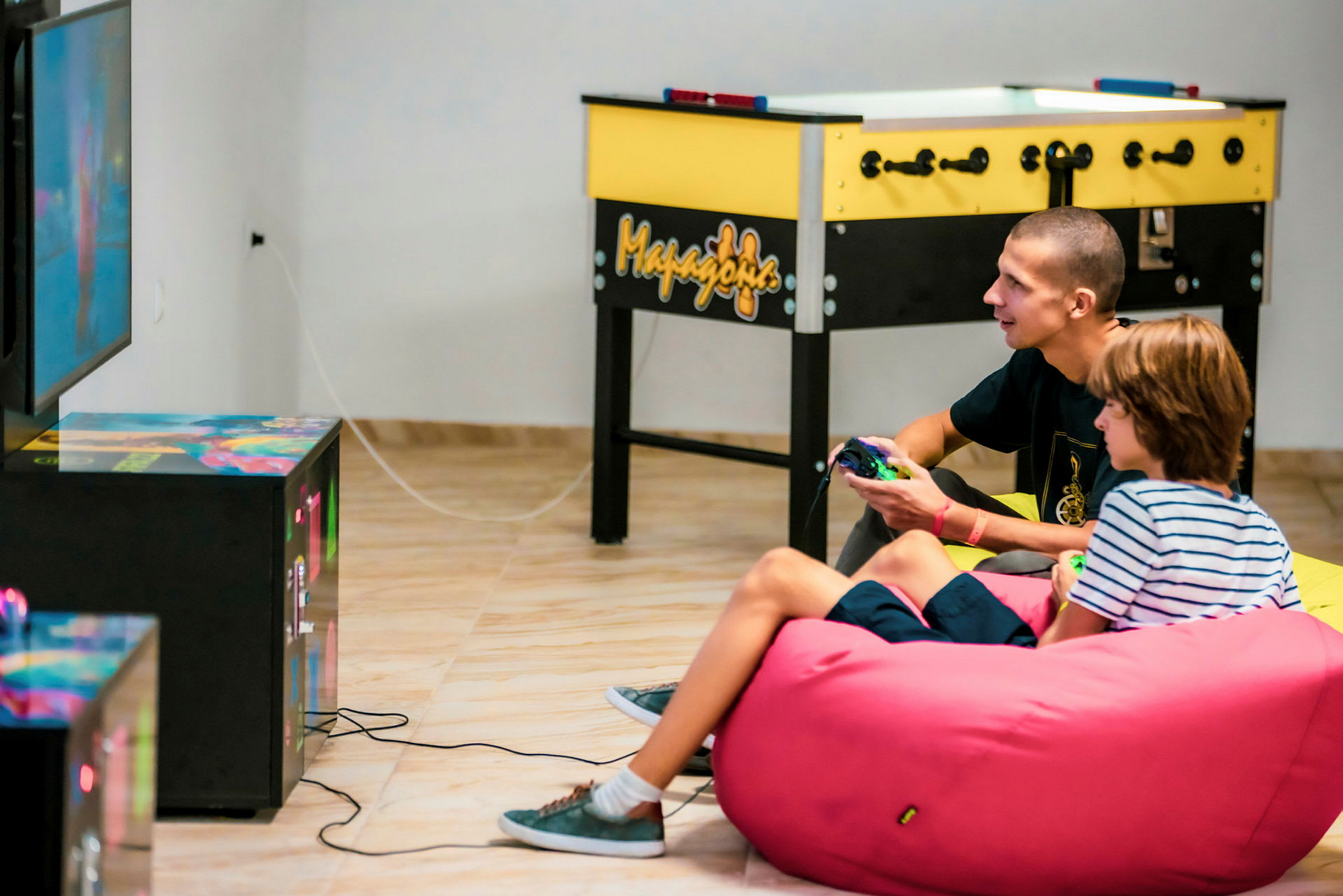 Två personer spelar TV-spel och sitter på en rosa sittsäck. I bakgrunden syns ett bordsfotbollsspel. (AI-genererad text)