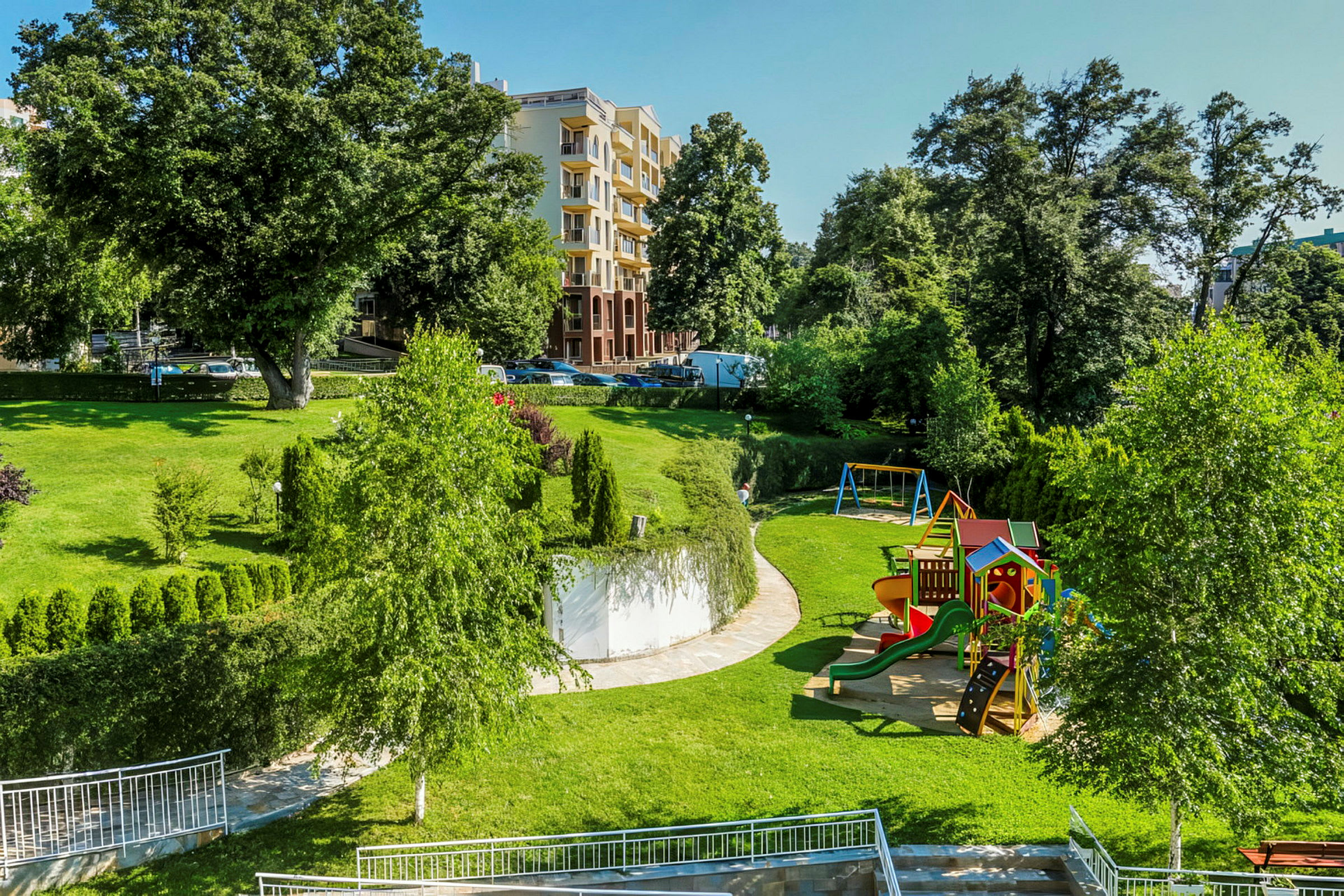 En park med lekplats, gräsmattor och träd. I bakgrunden finns byggnader. (AI-genererad text)