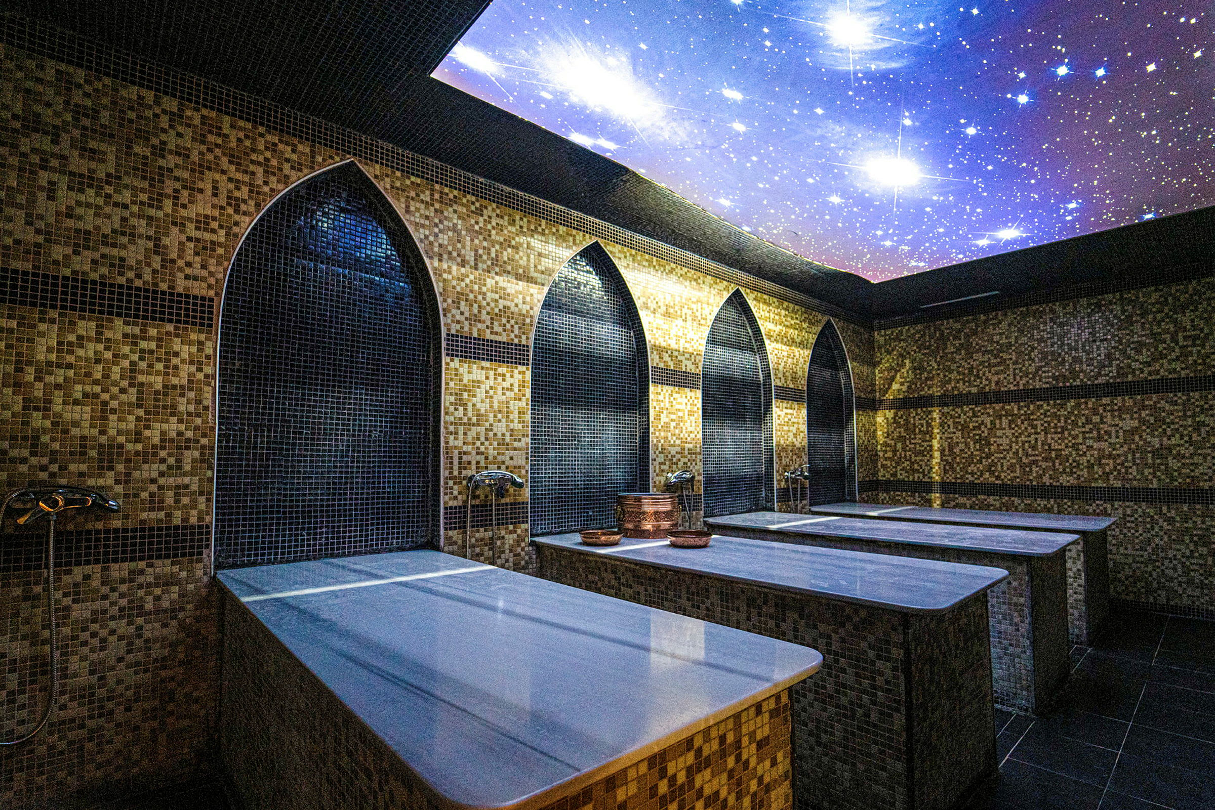 Rummet visar ett traditionellt hamam med marmorbänkar och dekorativ mosaik på väggarna. Taket är prytt med ett motiv av stjärnor. (AI-genererad text)