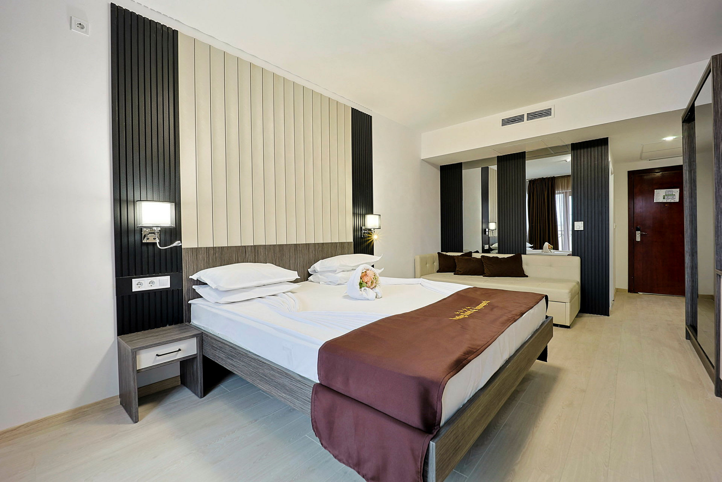 Ett hotellrum med en dubbelsäng, ljusa väggar och golv samt en brun dekorativ överkast. Rummet har en modern inredning med en sittplats och speglar. (AI-genererad text)