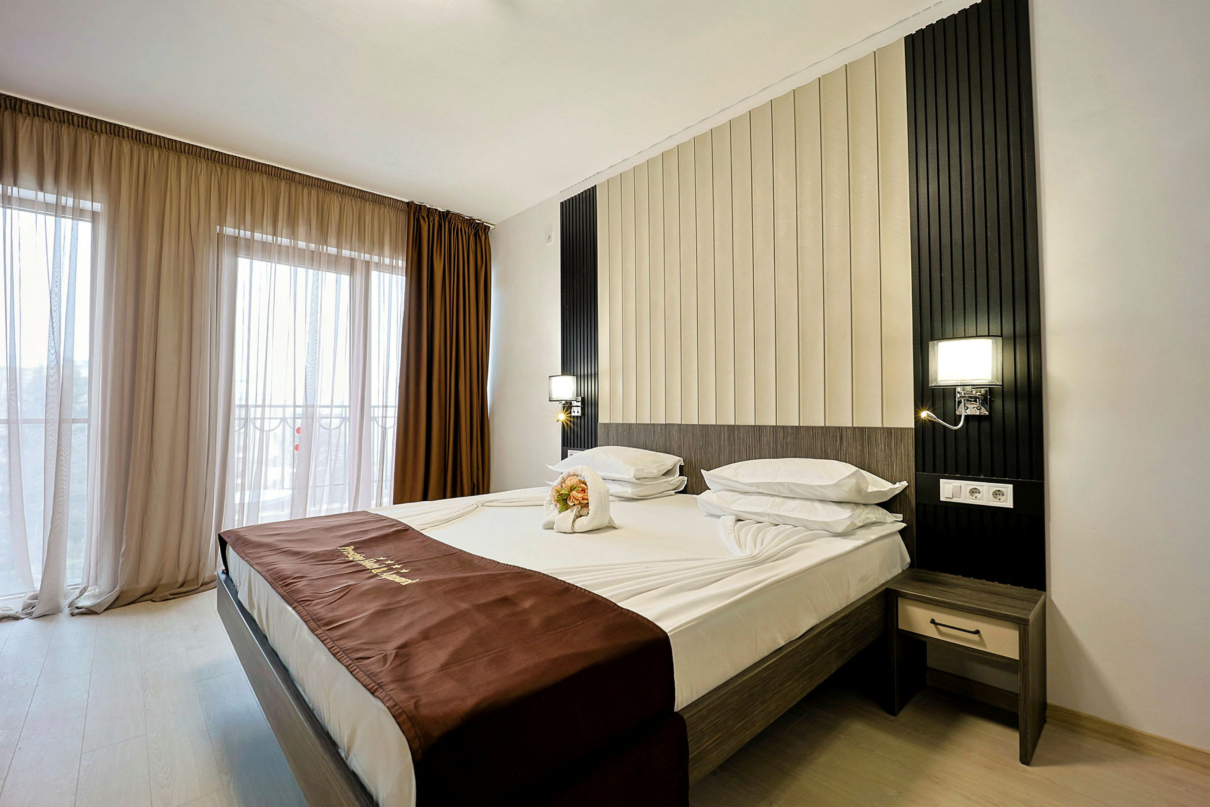 Ett modernt hotellrum med en bäddad dubbelsäng. Golvet är ljust och bakom sängen finns en dekorativ väggpanel. Fönstret är försett med skira och mörka gardiner. (AI-genererad text)