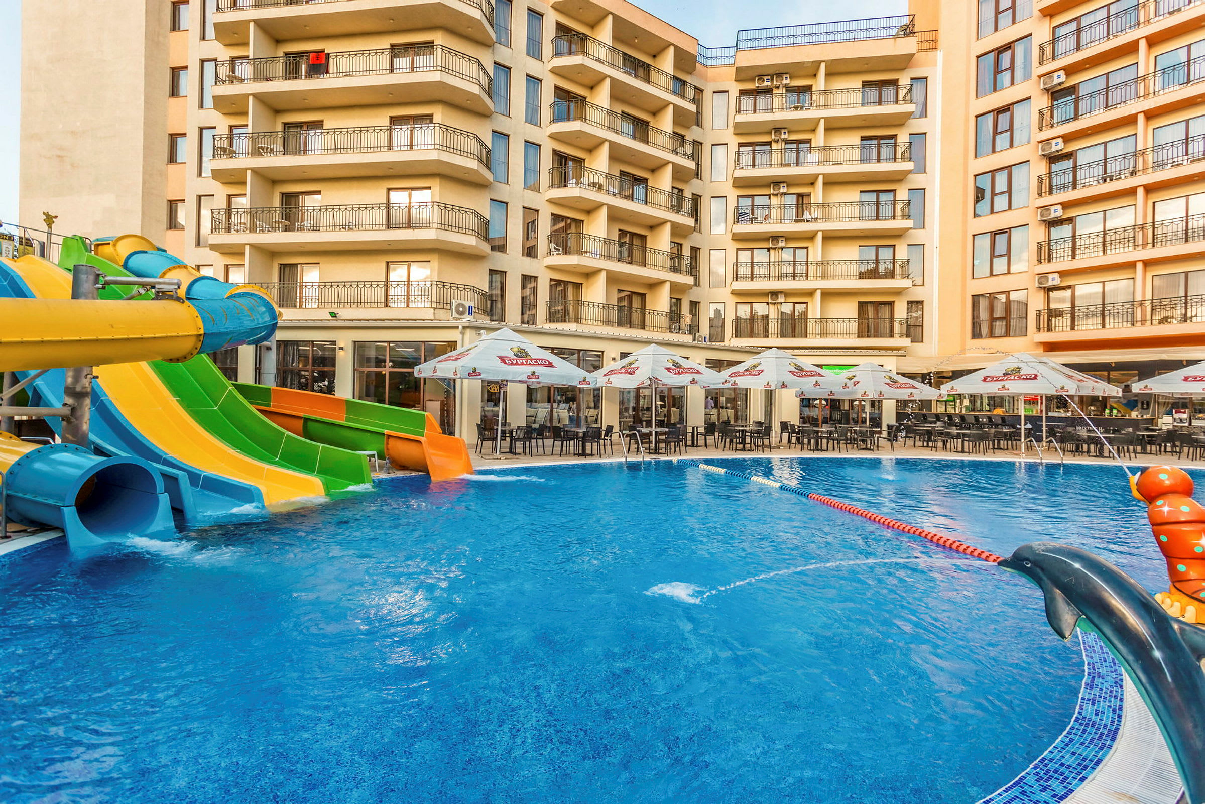 Bilden visar en swimmingpool med vattenrutschbanor i olika färger framför ett hotell, med utemöbler och parasoller vid poolområdet. (AI-genererad text)