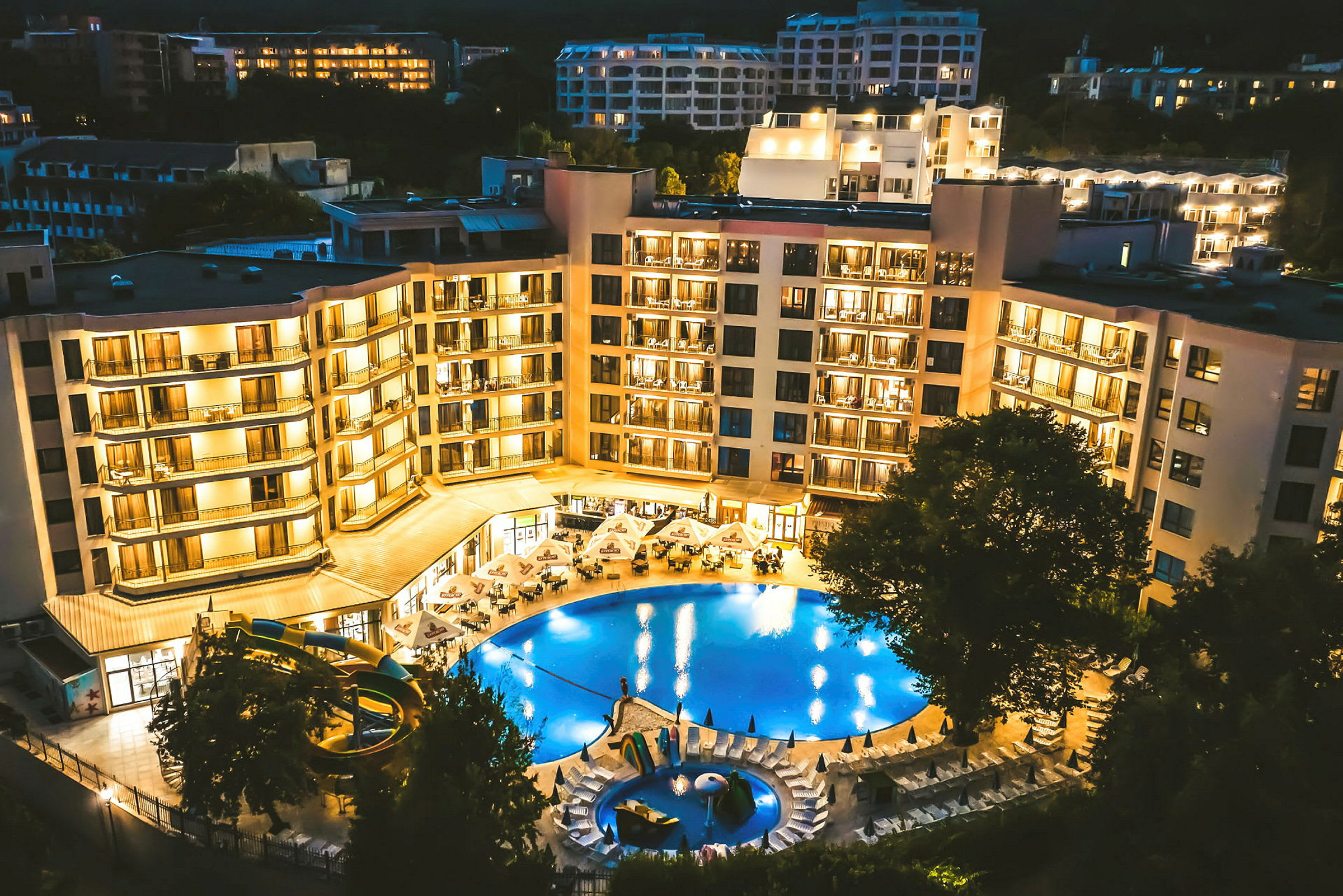 Upplyst hotellbyggnad med en pool och omgivande solstolar, fotograferad på kvällen. (AI-genererad text)