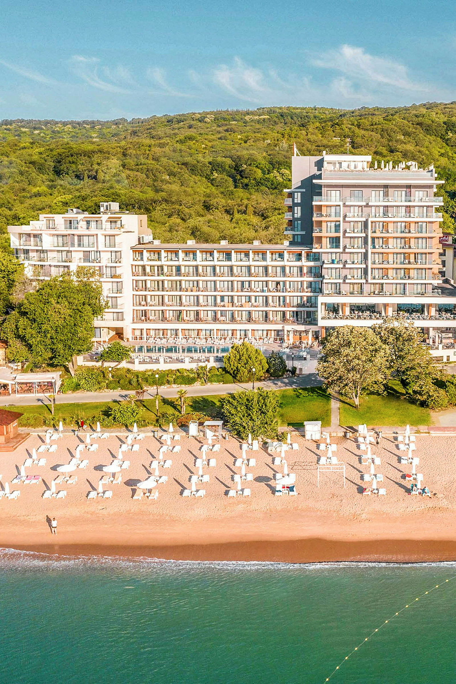 Vy över en strand med solstolar och parasoll vid ett hotell, omgiven av grönska. (AI-genererad text)