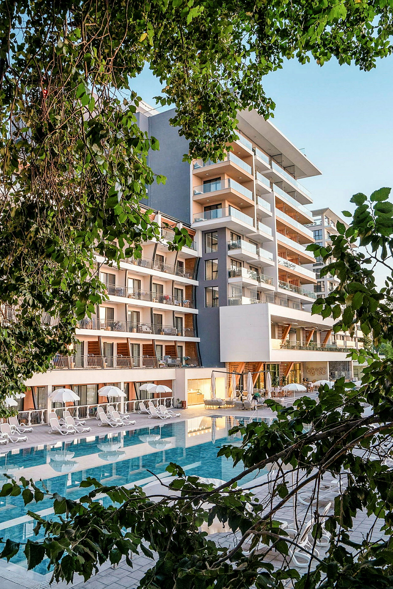 Hotellbyggnad med pool i förgrunden, omgiven av grönskande träd. (AI-genererad text)