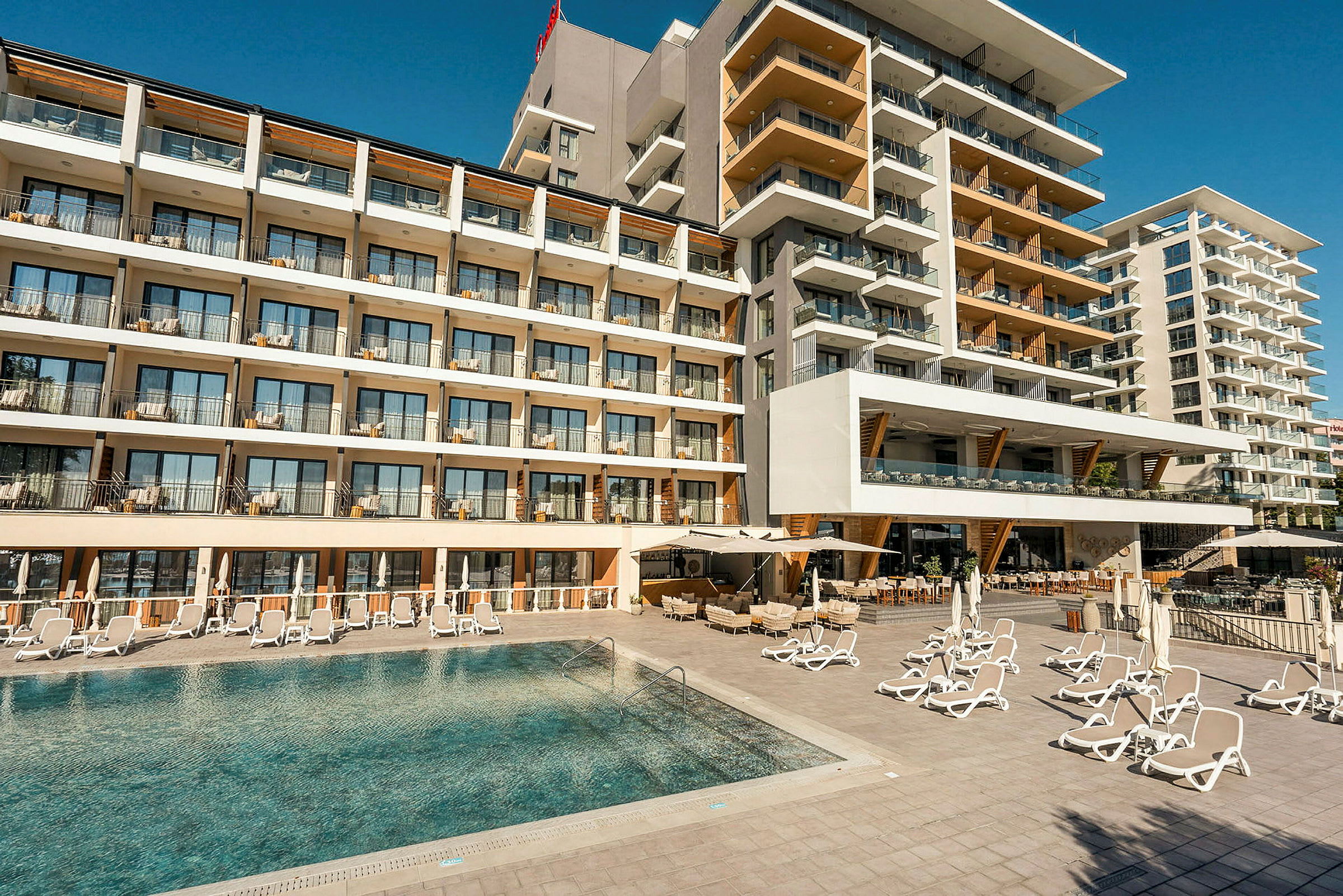 Hotellfasad med flera våningar och balkonger, framför en pool med solstolar. (AI-genererad text)